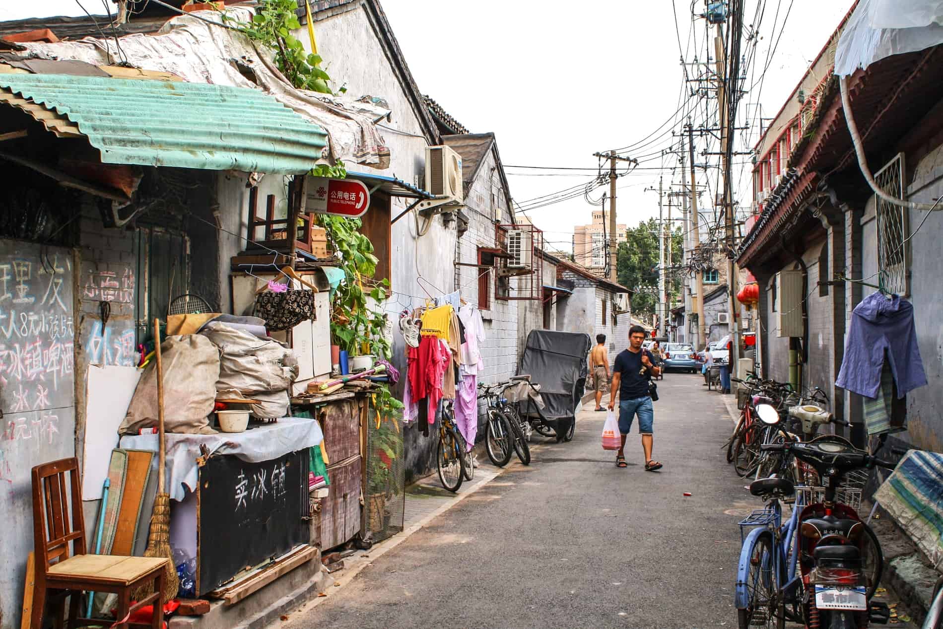 A man walks through a Beijing hutong street, amongst furniture, hanging laundry and bikes. 