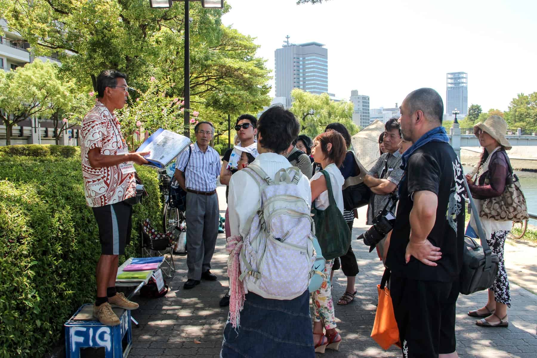 A peace activist in Hiroshima stands on a bench and talks to a group of visitors. 