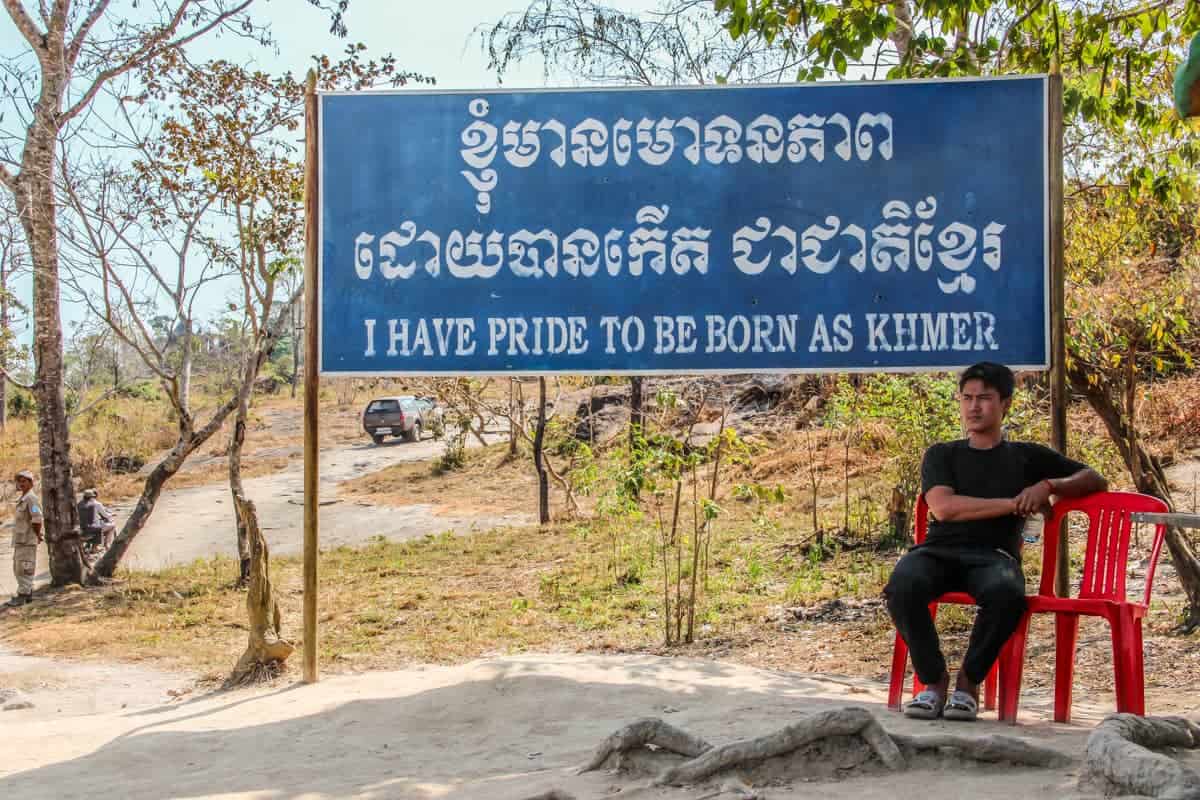 A man sits next to a blue sign saying 'I have pride to be born as Khmer' at the Preah Vihear temple entrance