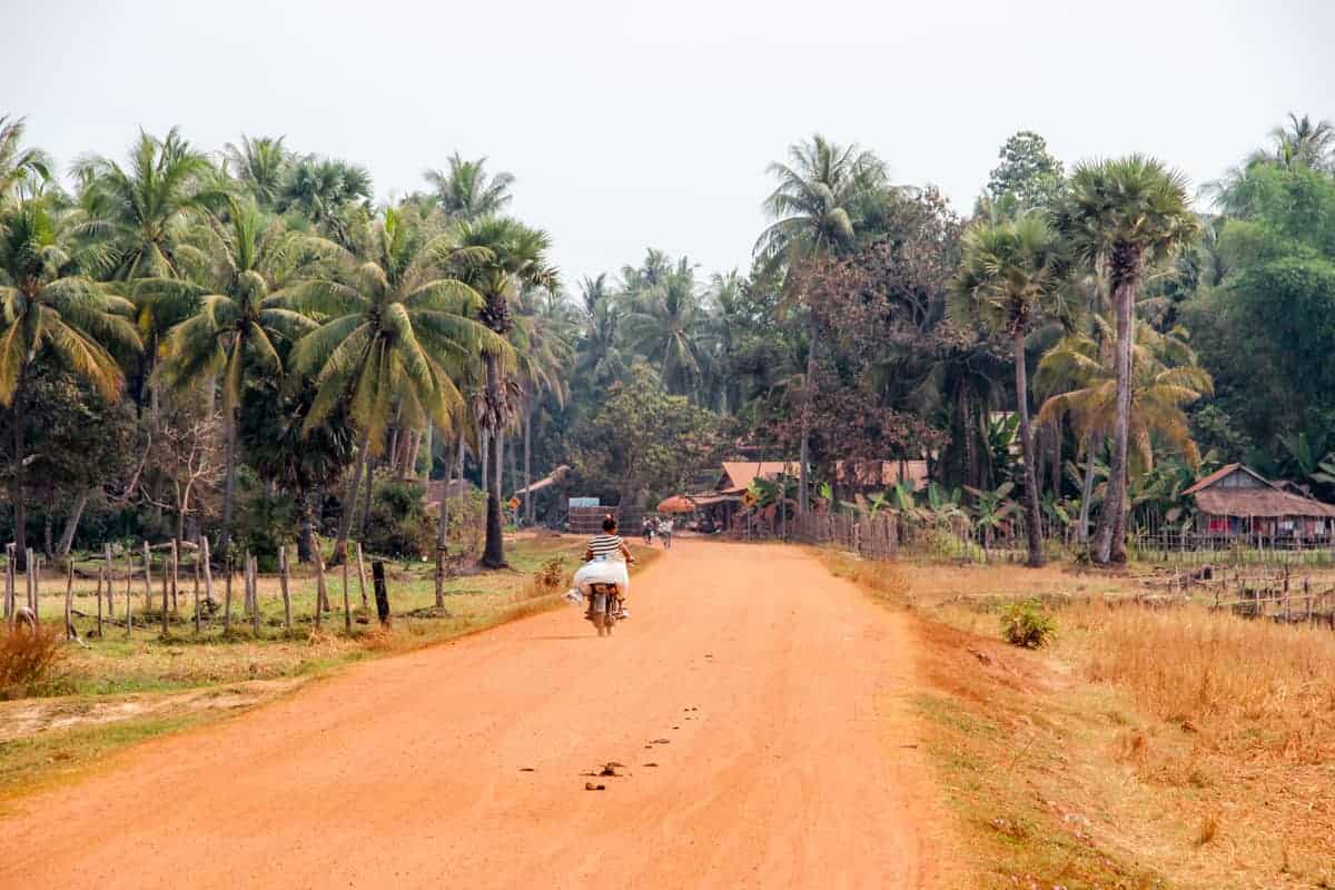 A long and wide orange dusty road leading to a small village of wooden houses in Cambodia