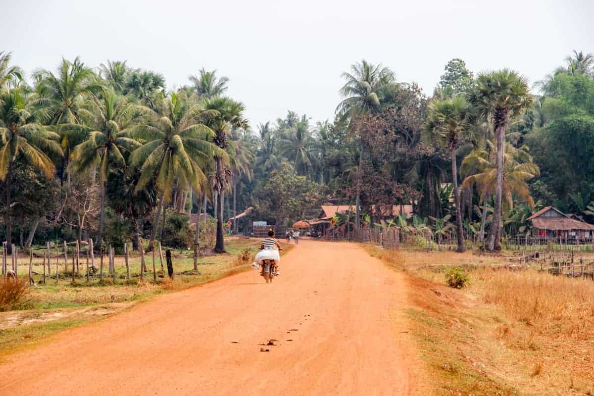 Un largo y ancho camino polvoriento naranja que conduce a un pequeño pueblo de casas de madera en Camboya