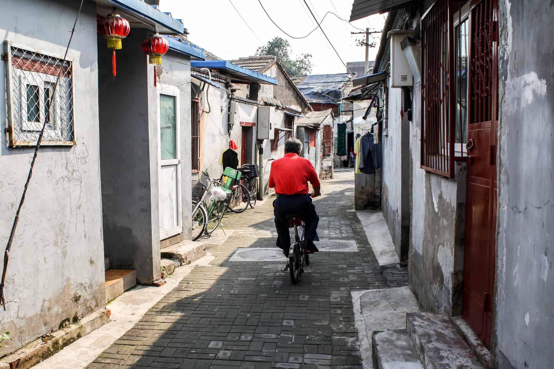 A man in a red shirt riding a bicycle on a narrow street in a residential Beijing Hutong. 
