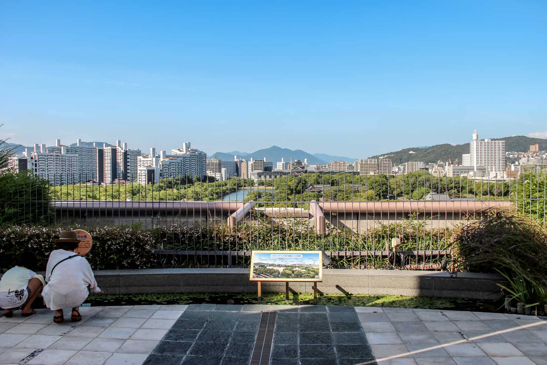 Tourists bend down to look at a sign at a viewpoint over Hiroshima city's modern skyline, Castle and gardens.