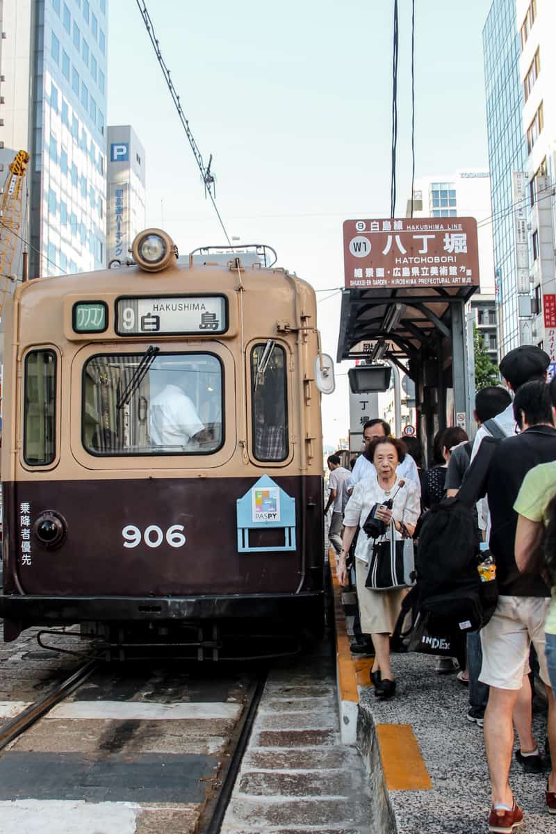 A crowd of people on the platform next to a brown tram in Hiroshima. 
