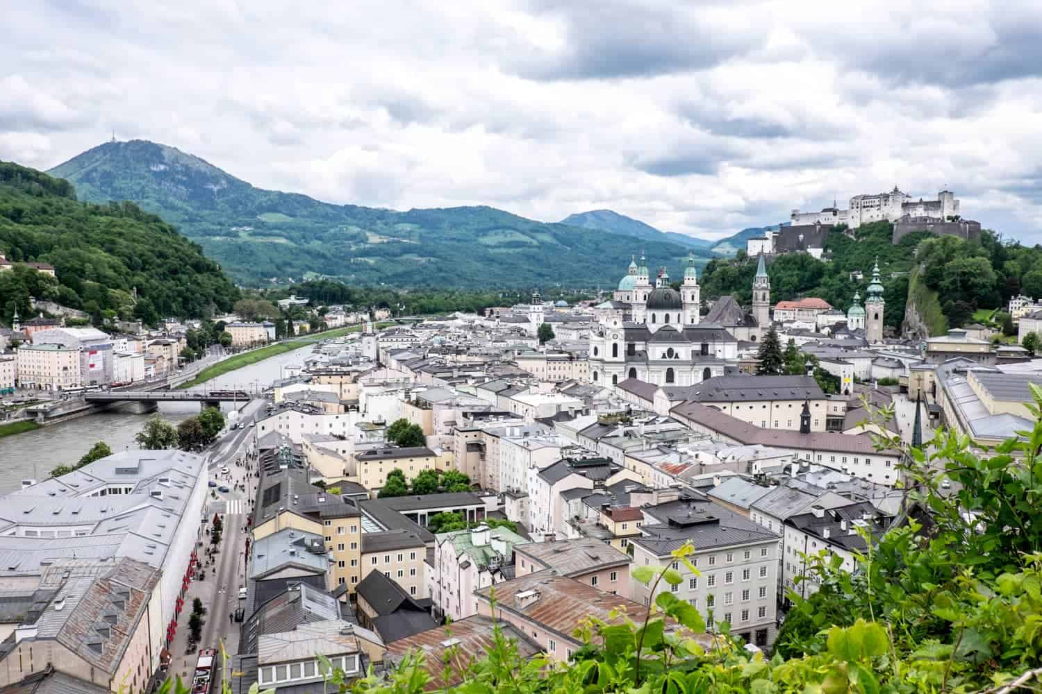 Elevated view of Salzburg - an old riverside town with a castle on the hill backed by forested mountains. 