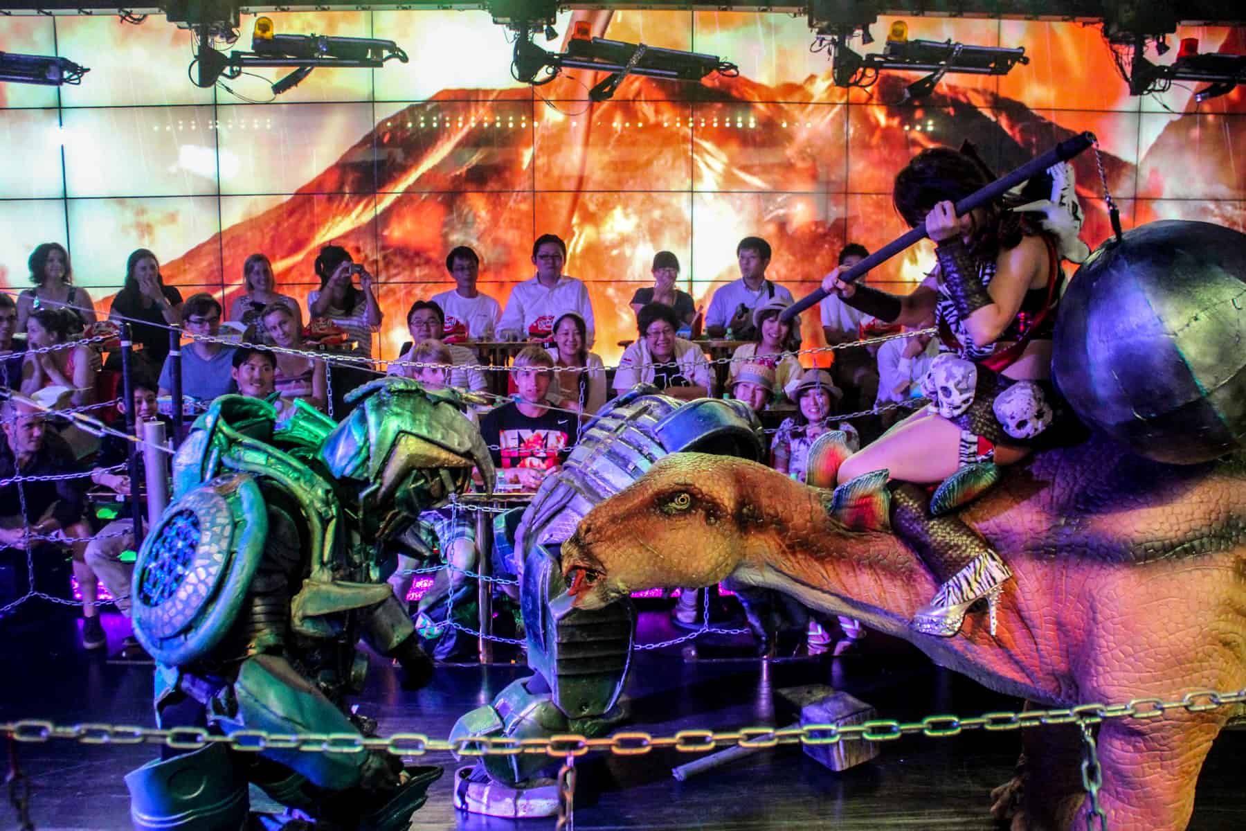 The audience looks on as a woman riding a dinosaur tries to hit a robot with a giant ball at the Robot show in Tokyo.