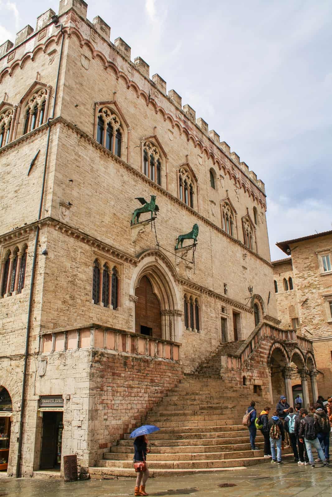 A walking tour in Perugia gathers outside of a gothic palace with green lion and dragon sculptures above its archway. 