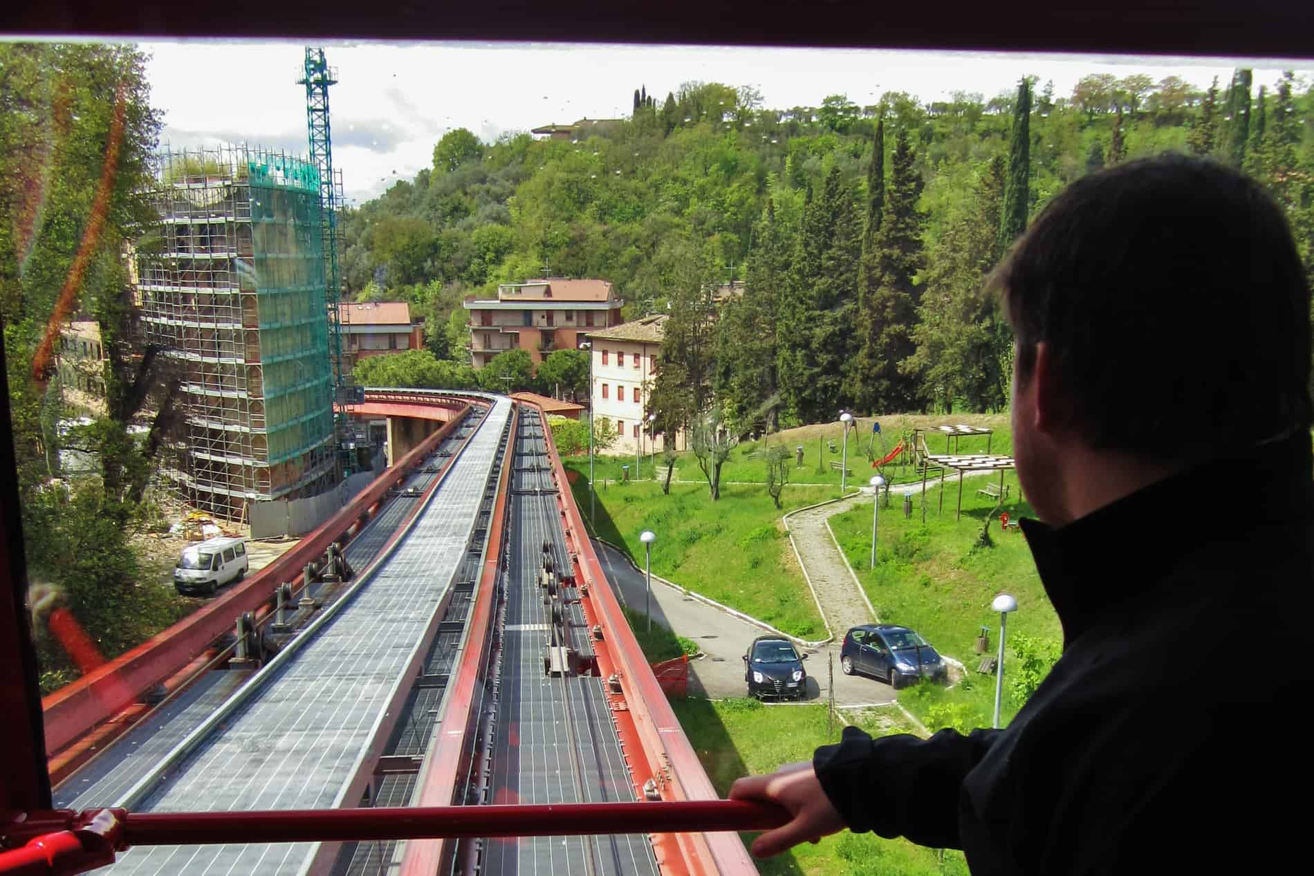A man looks out the widnow of the Perugia Minimetro with a view to the small track curving through a forest. 