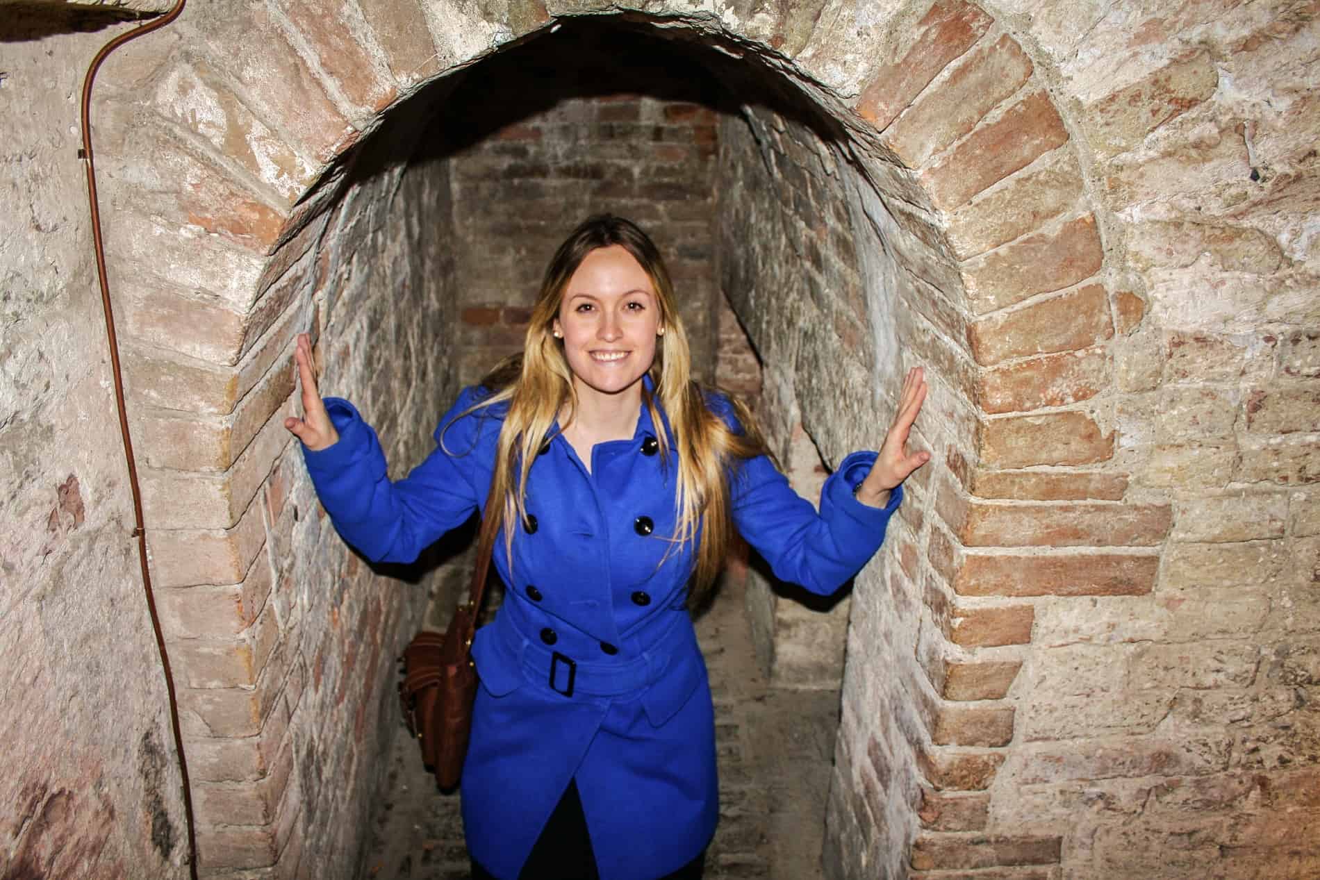 A woman in a blue coat crouches in the brick archway of an underground tunnel in the Rocca Paolina Fortress in Perugia. 