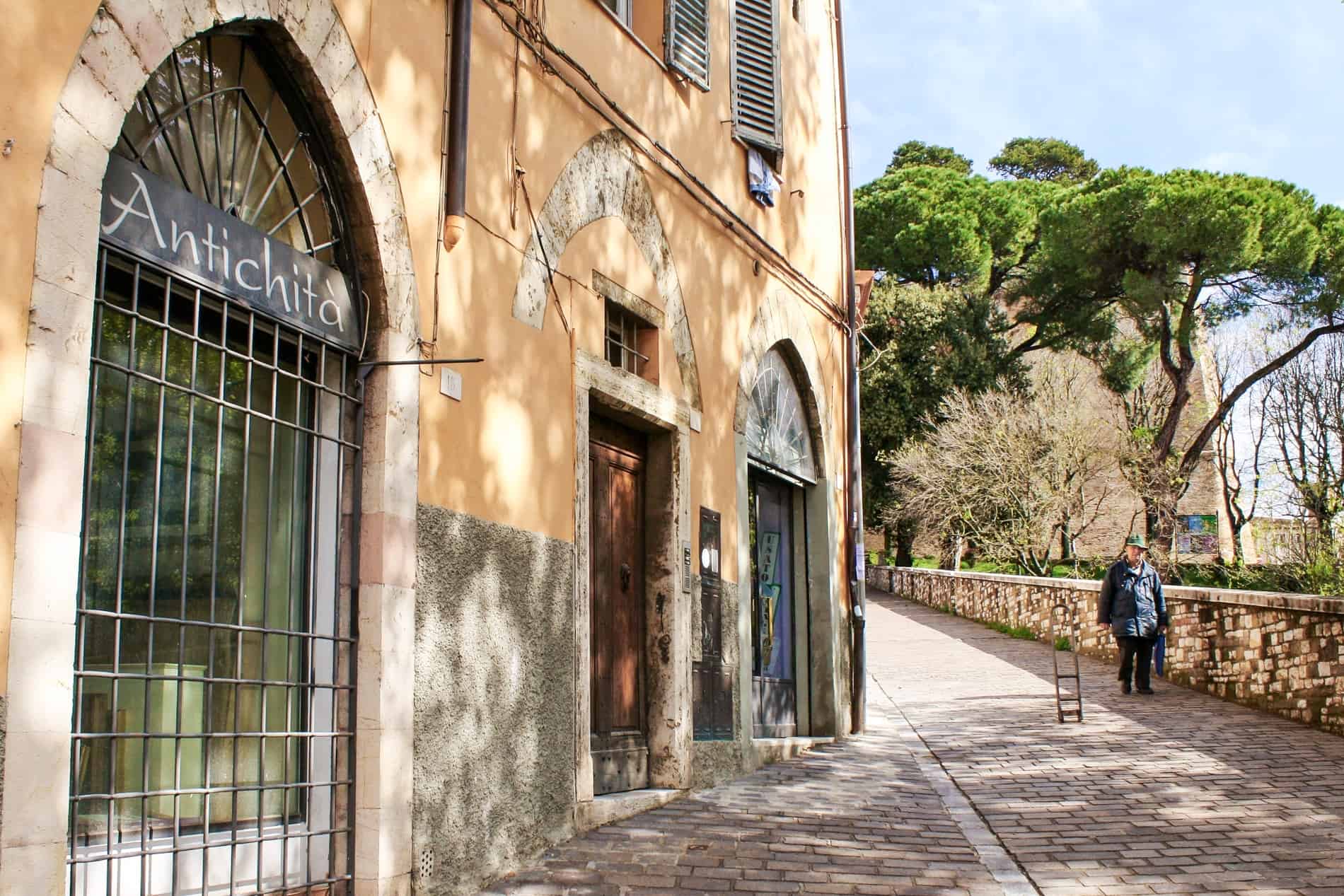 A person walks down a paved, sloped path next to a sandstone building with gated archways. 