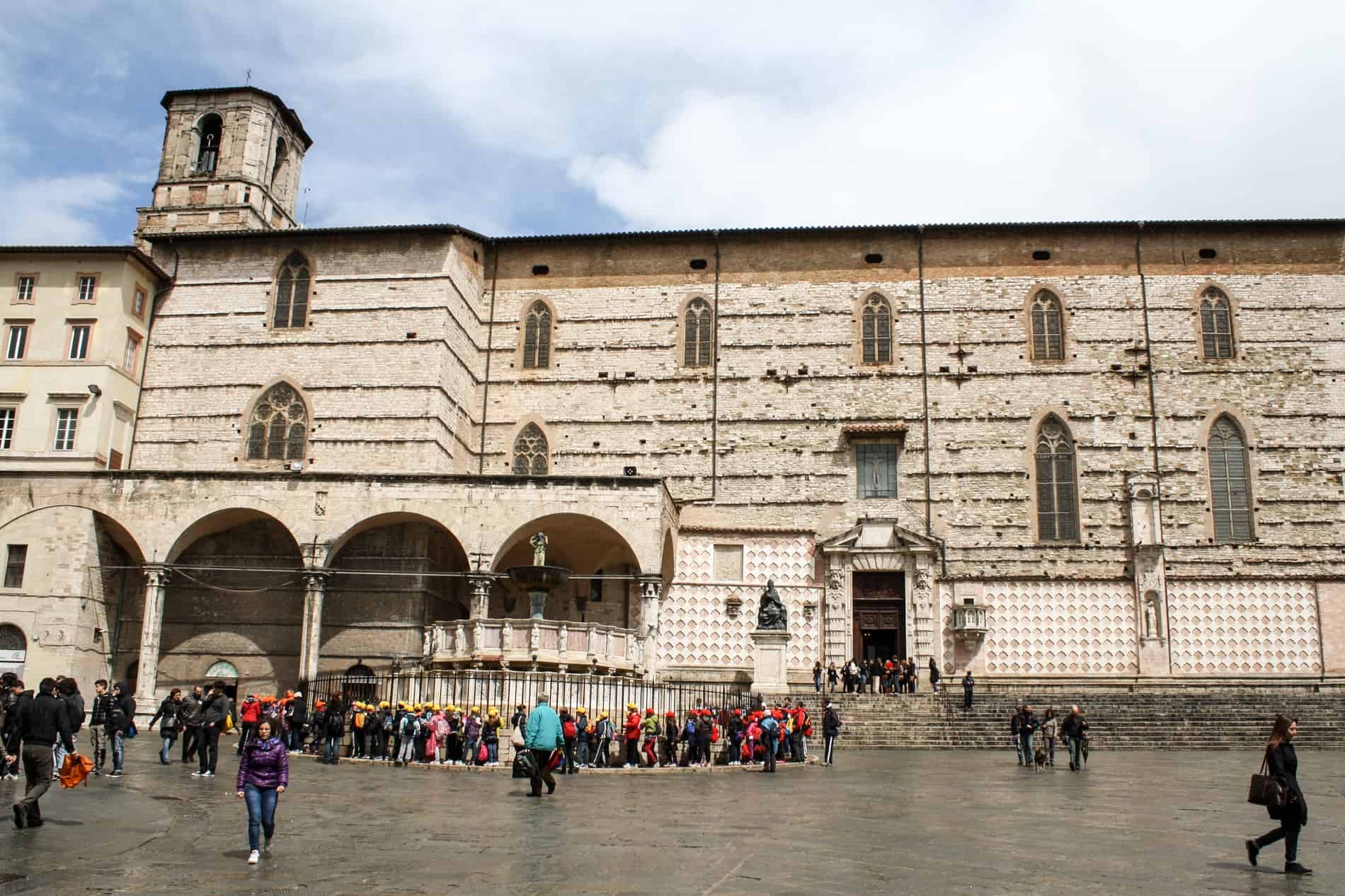 Groups of tourists gather around the marble Fontana Maggiore in front of the huge stone landmark of the Perugia Cathedral.