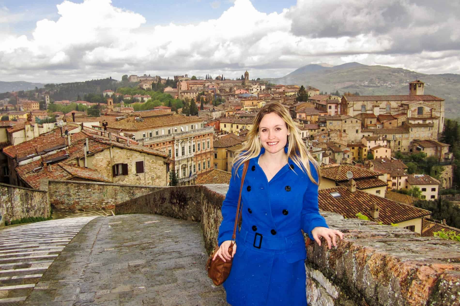 A woman in a blue coat standing on a stone bridge to the golden backdrop of Perugia city on a hill. 