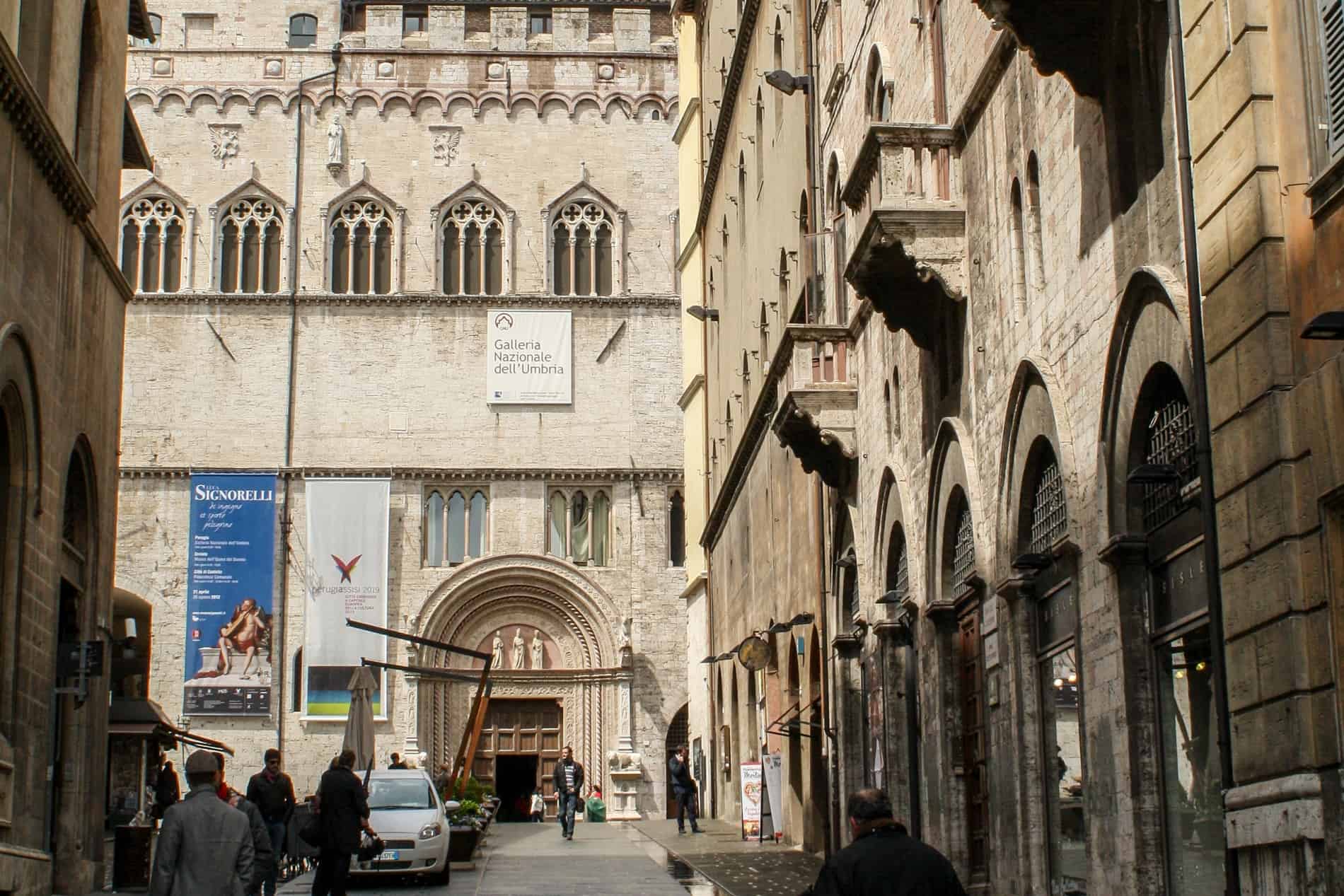 A street of light golden stone structures leading to the gothic building with a textile sign for the Galleria Nazionale dell'Umbria (The National Gallery of Umbria).