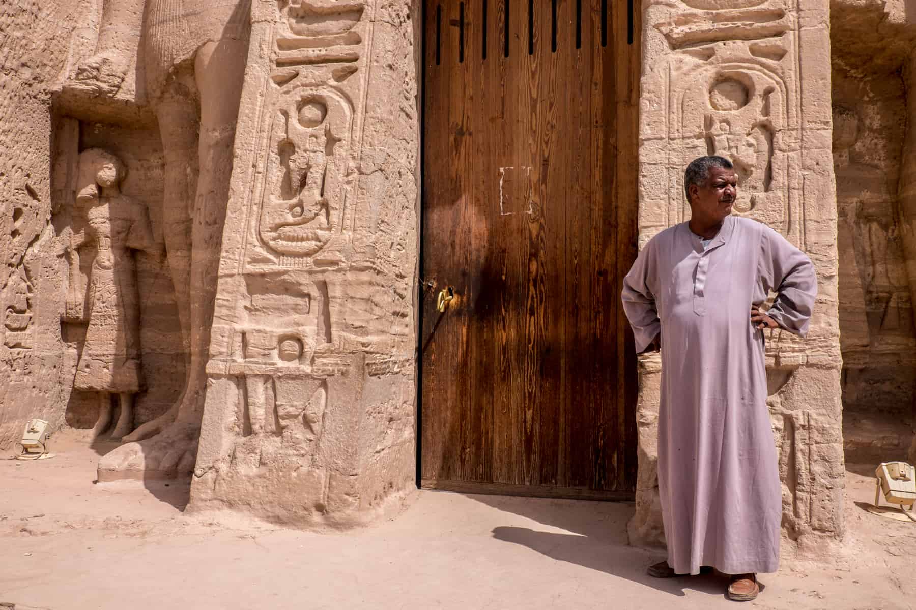 An Egyptian man wearing a long tunic guards a wooden entrance door to the rock carved Abu Simbel temple near Aswan, Egypt