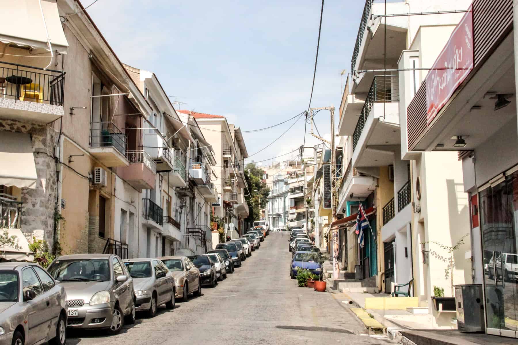 Streetview of a coastal neighbourhood in Athens lined with low-rise beige and white block buildings and cars. 