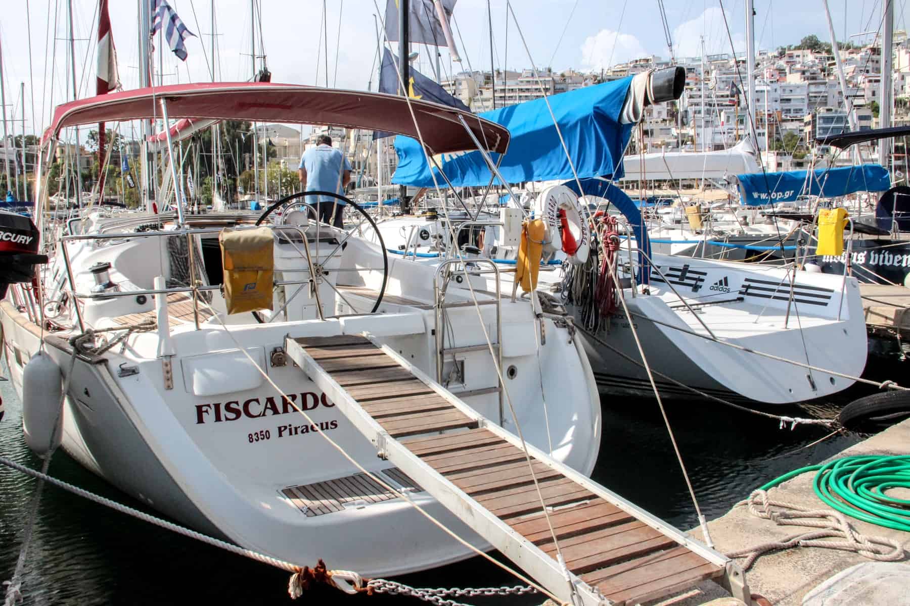 A man in a blue t-shirt stands on his boat 'Fiscardo' in a marina lined with boats in front of a bay of apartment buildings. 