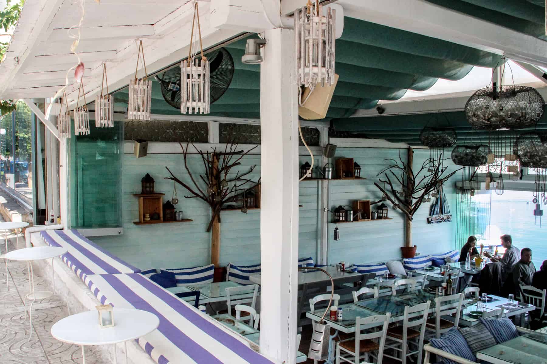 People dining in a teal painted restaurant with blue and white stripped cushions, that overlooks the sea. 