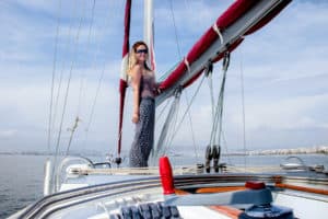 A woman standing on the bow of a yacht, holding onto a rolled up red sail. In the distance is the Athens coastline.