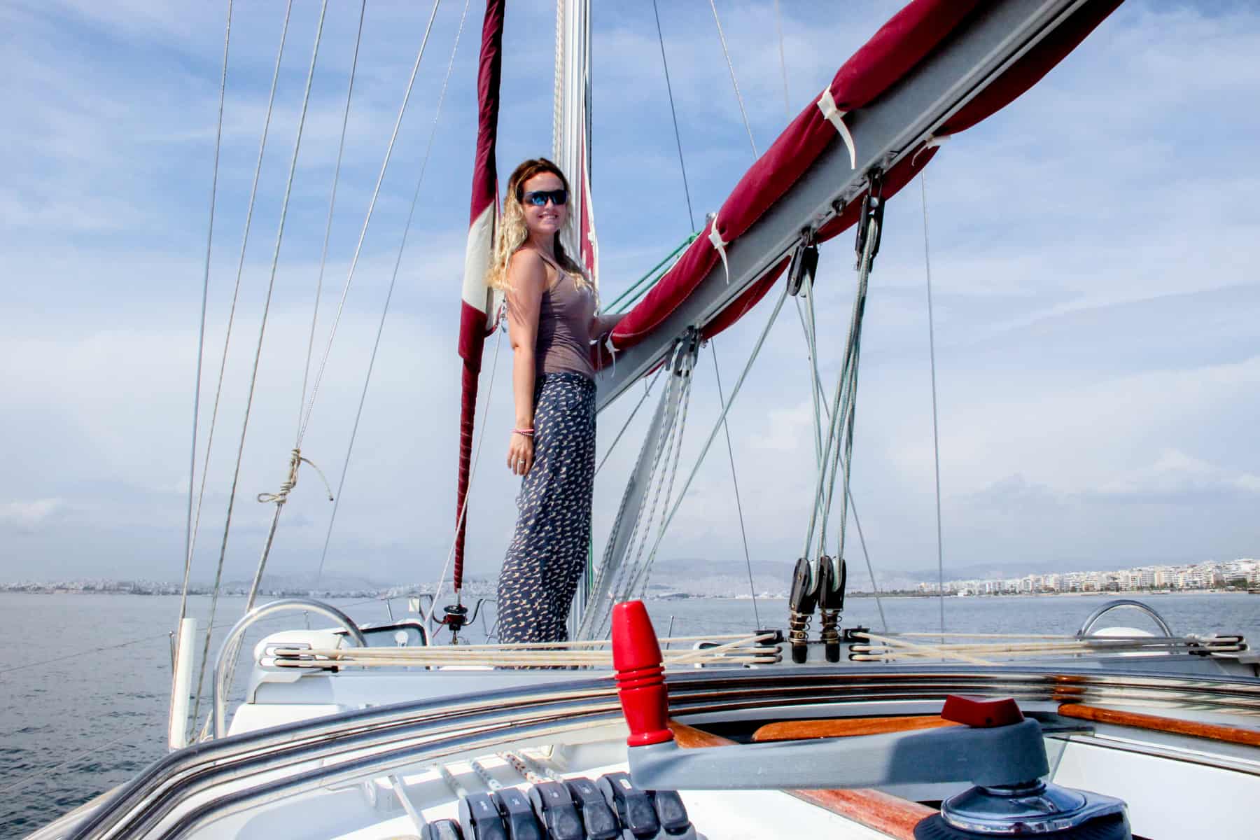A woman standing on the bow of a yacht, holding onto a rolled up red sail. In the distance is the Athens coastline. 