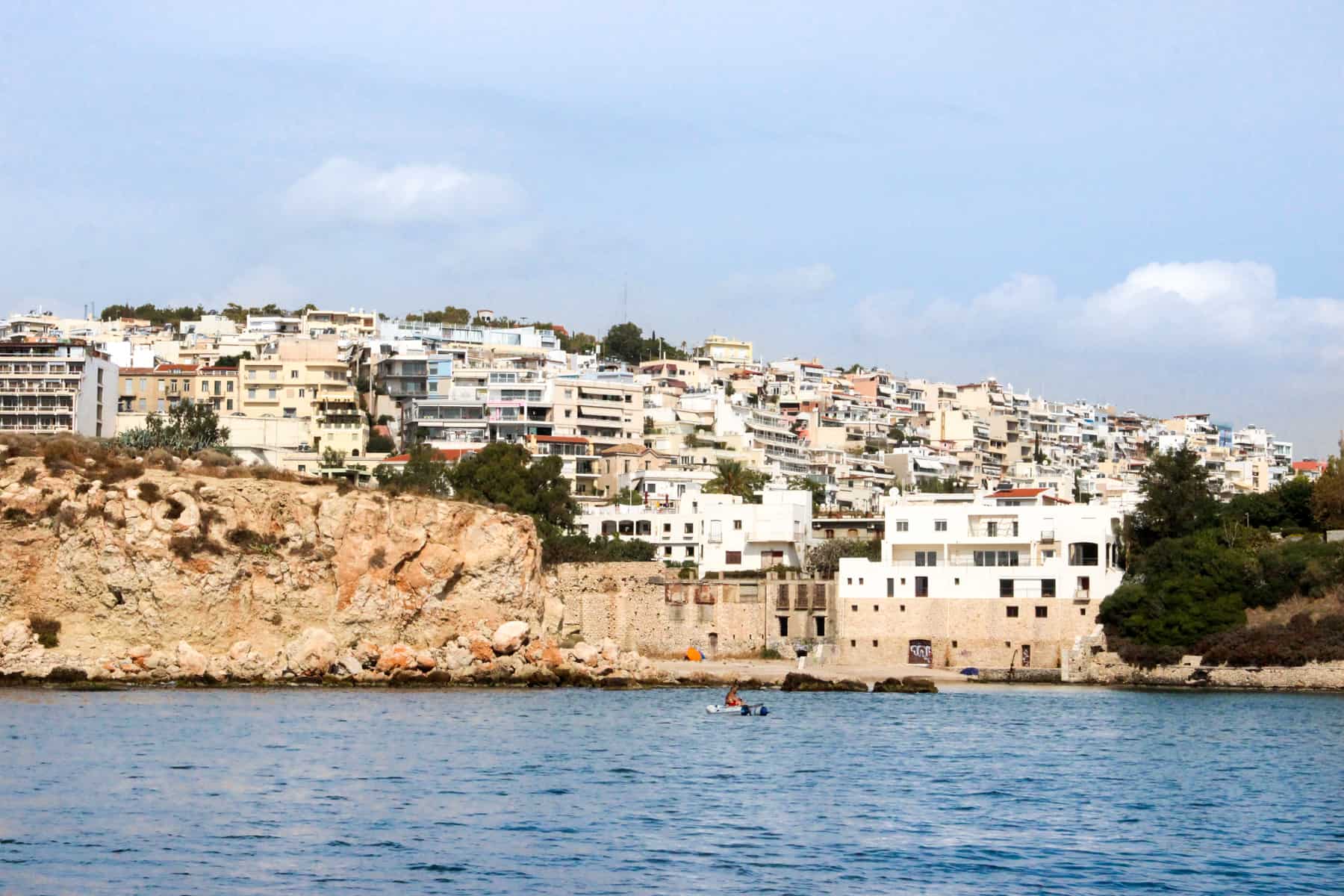 A man in a tiny boat floating on the blue waters in front of a rock wall and a hill of apartment buildings. 