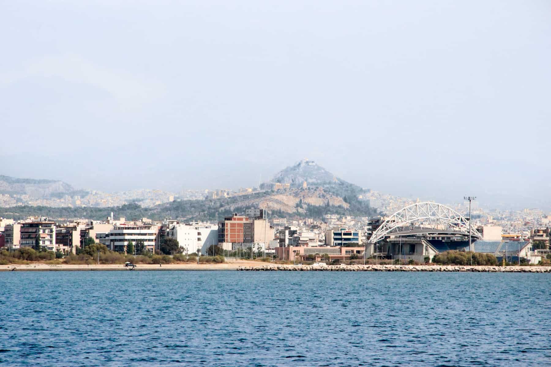 View from the sea of the Athens Riviera coastline filled with apartment buildings and a stadium with one of the city hills in the background.