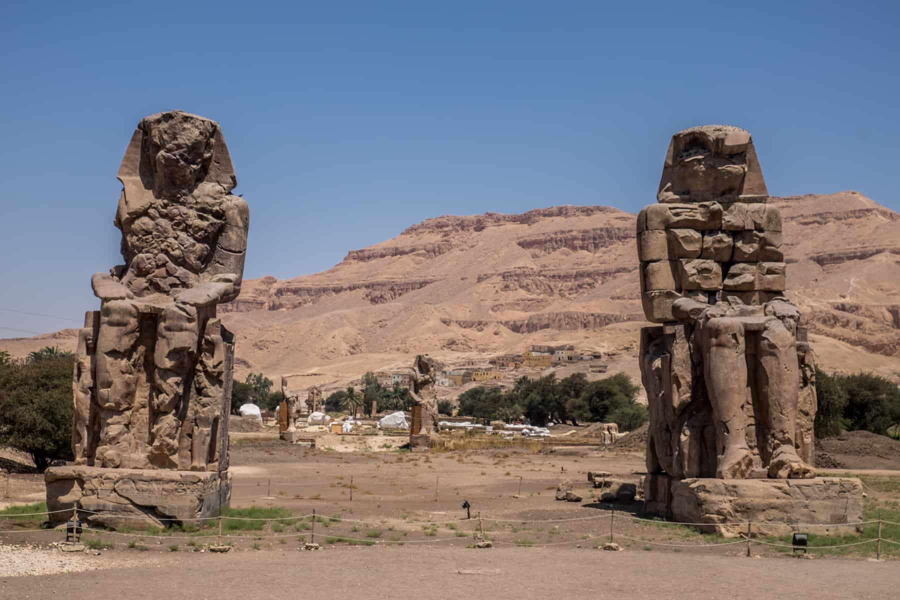 Three large statues of the Colossi of Memnon stand on a patch of grass in front of mountains in Luxor, Egypt