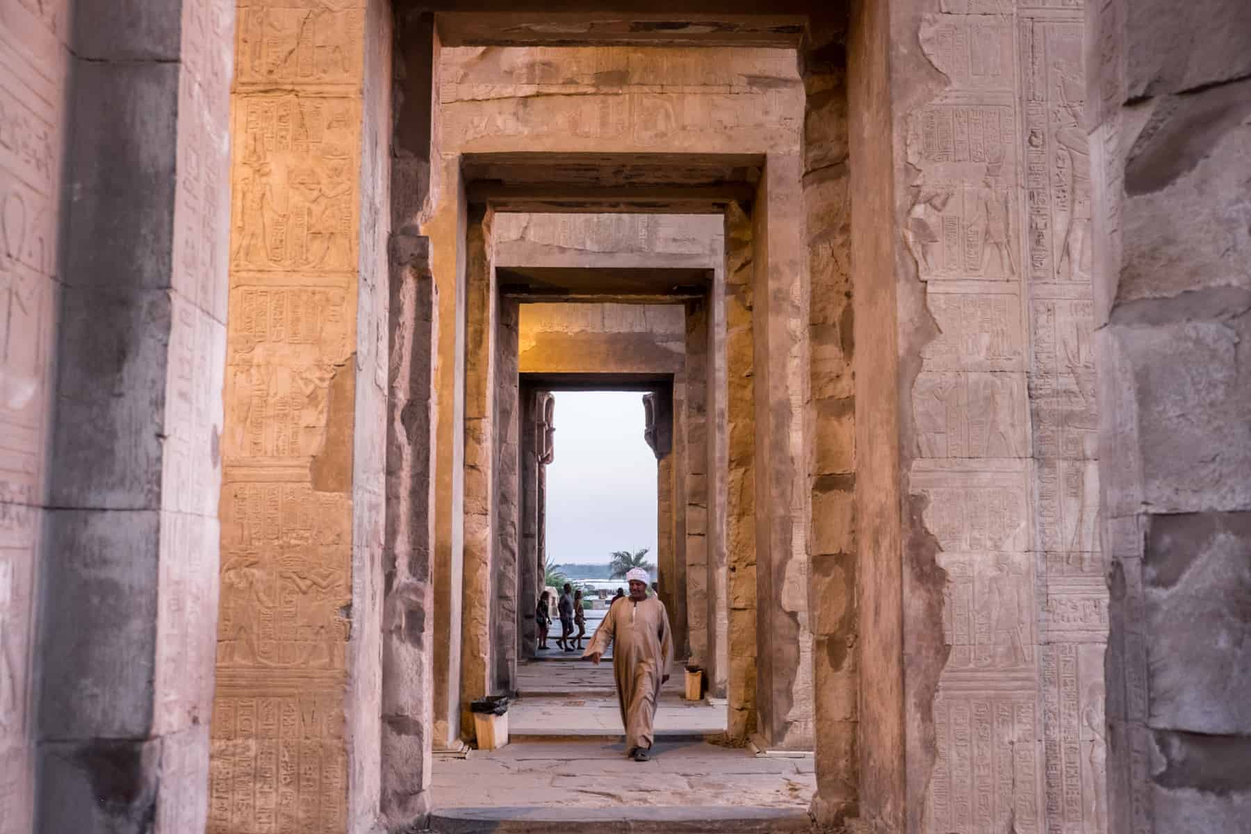 An Egyptian Guard in a white tunic walks through the square stoned corridor of Kom Ombo temple at sunset, that sets the temple in an orange glow
