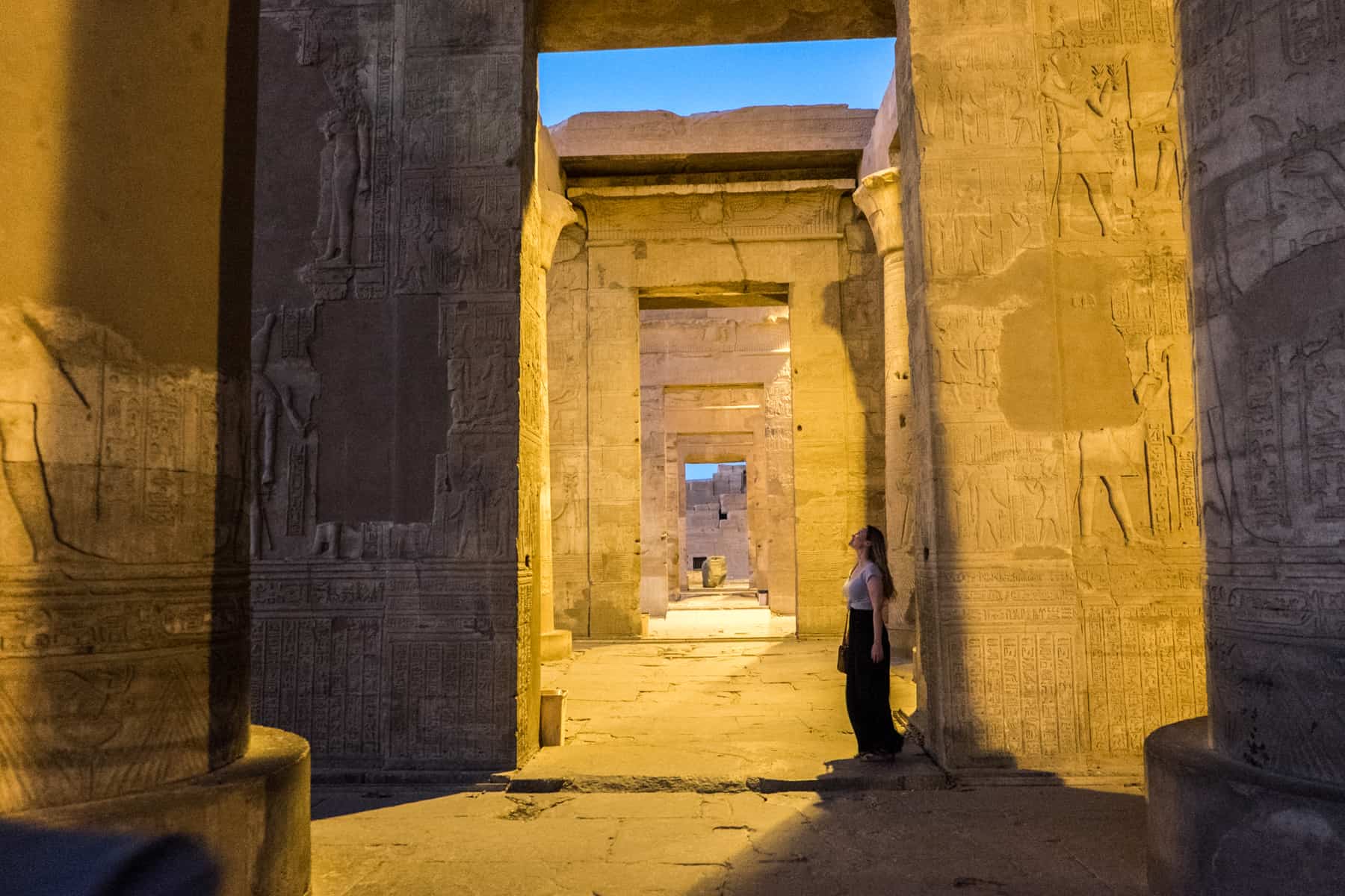 A woman stands and stares up at the tall walls of carvings at the Kom Ombo temple in Egypt, glowing in yellow light when visited in the evening