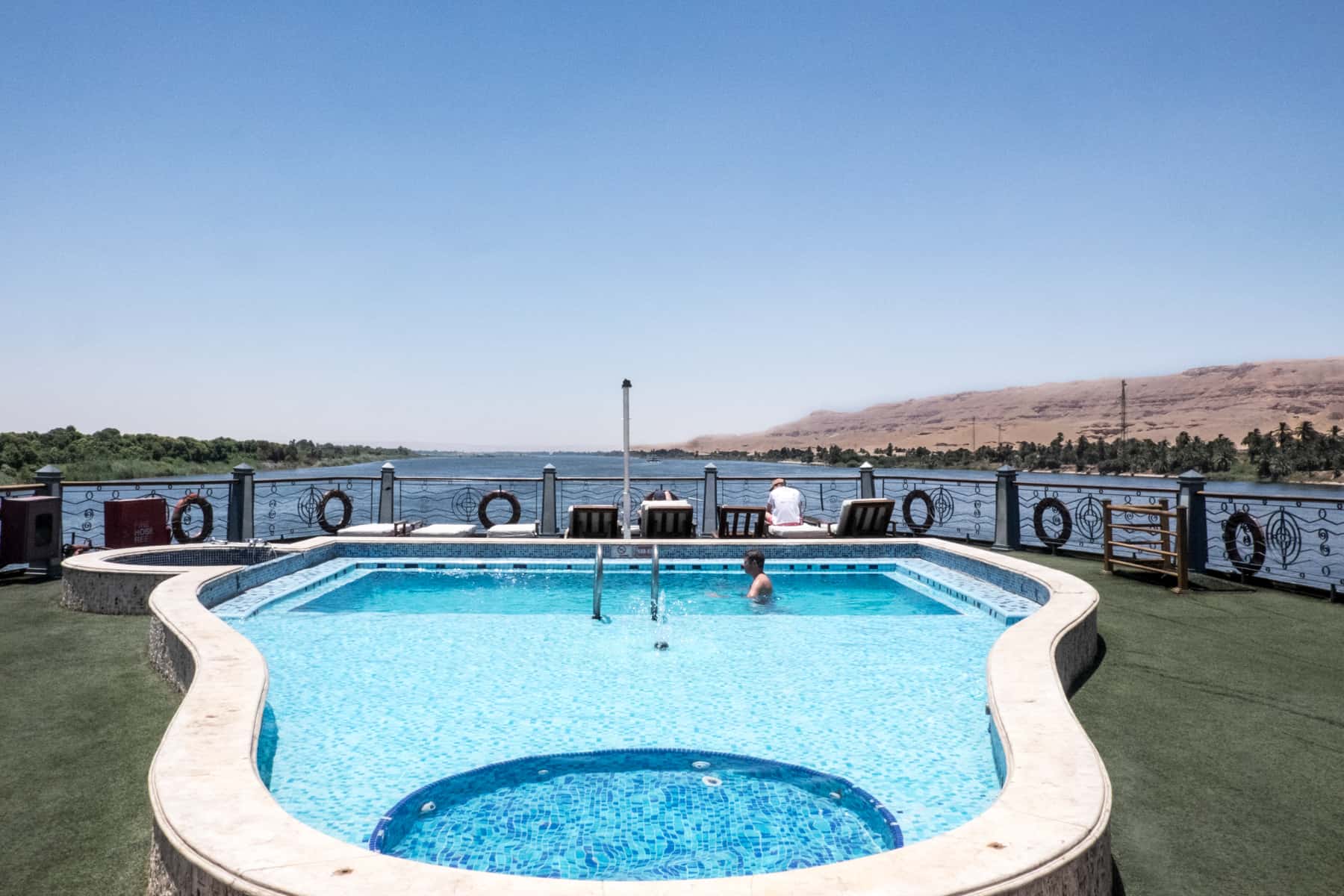 A swimming pool on the top deck of a nile cruise boat in Egypt, with the Nile River rock formations in the background