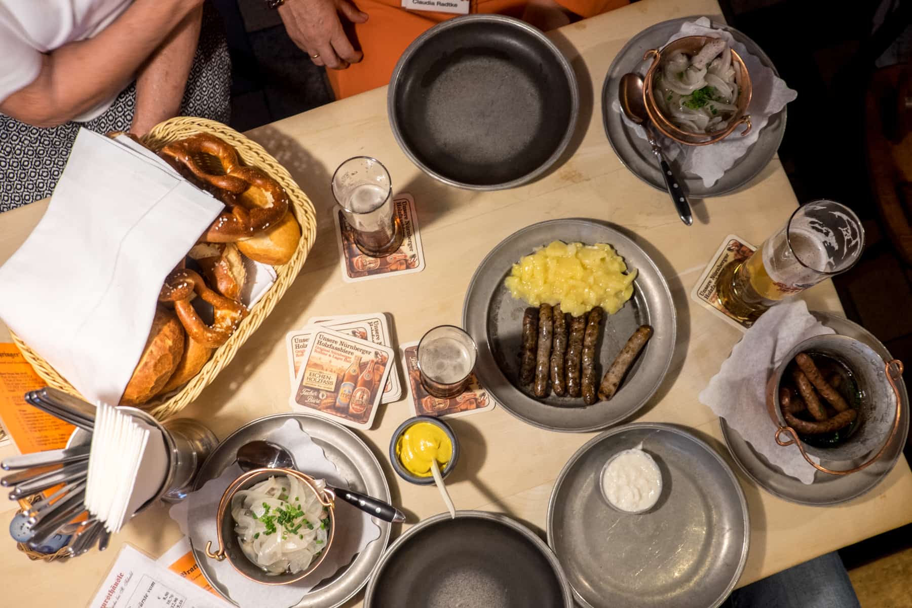 Vista de Ariel de una mesa en una taberna de Nuremberg con cuatro platos de salchichas, cerveza, cubiertos y pan.