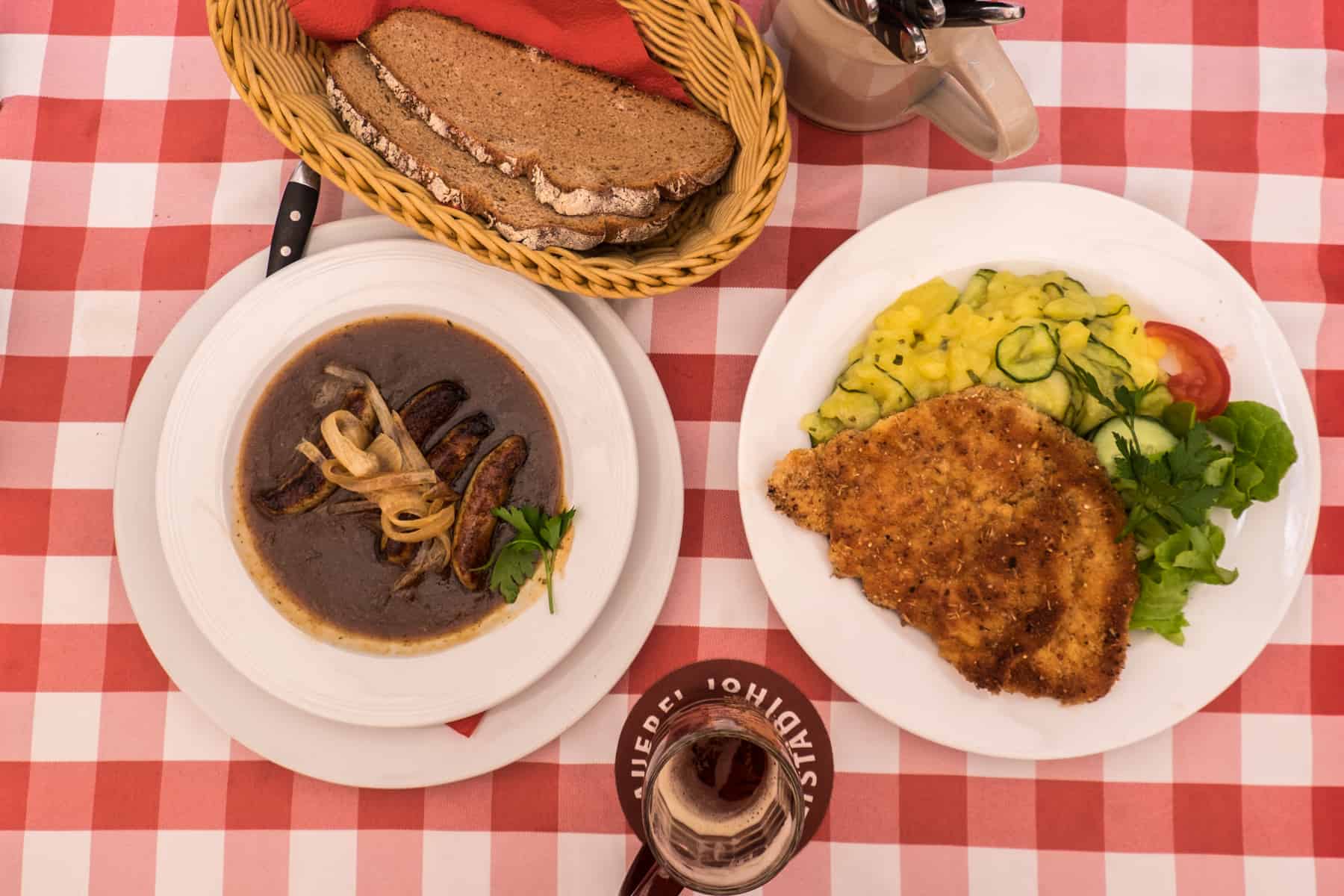 A red and white checkered tablecloth full of Nuremberg food specialities -beer battered schnitzel, a dish of nuremberg sausages in gravy, alongside a basket of bread and a beer