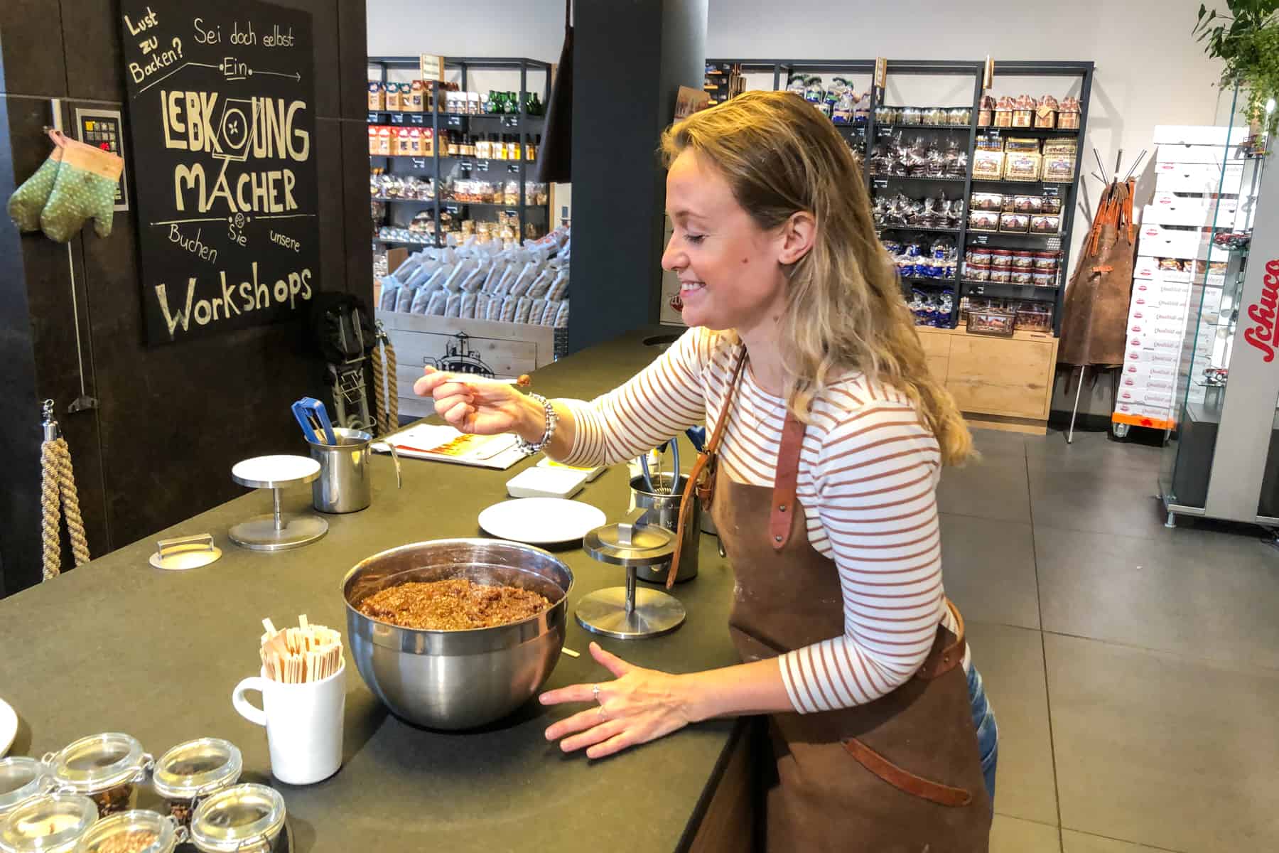 A woman in a striped top and brown leather apron takes a spoonful of Nuremberg gingerbread mixture from a large metal bowl and is about to eat it