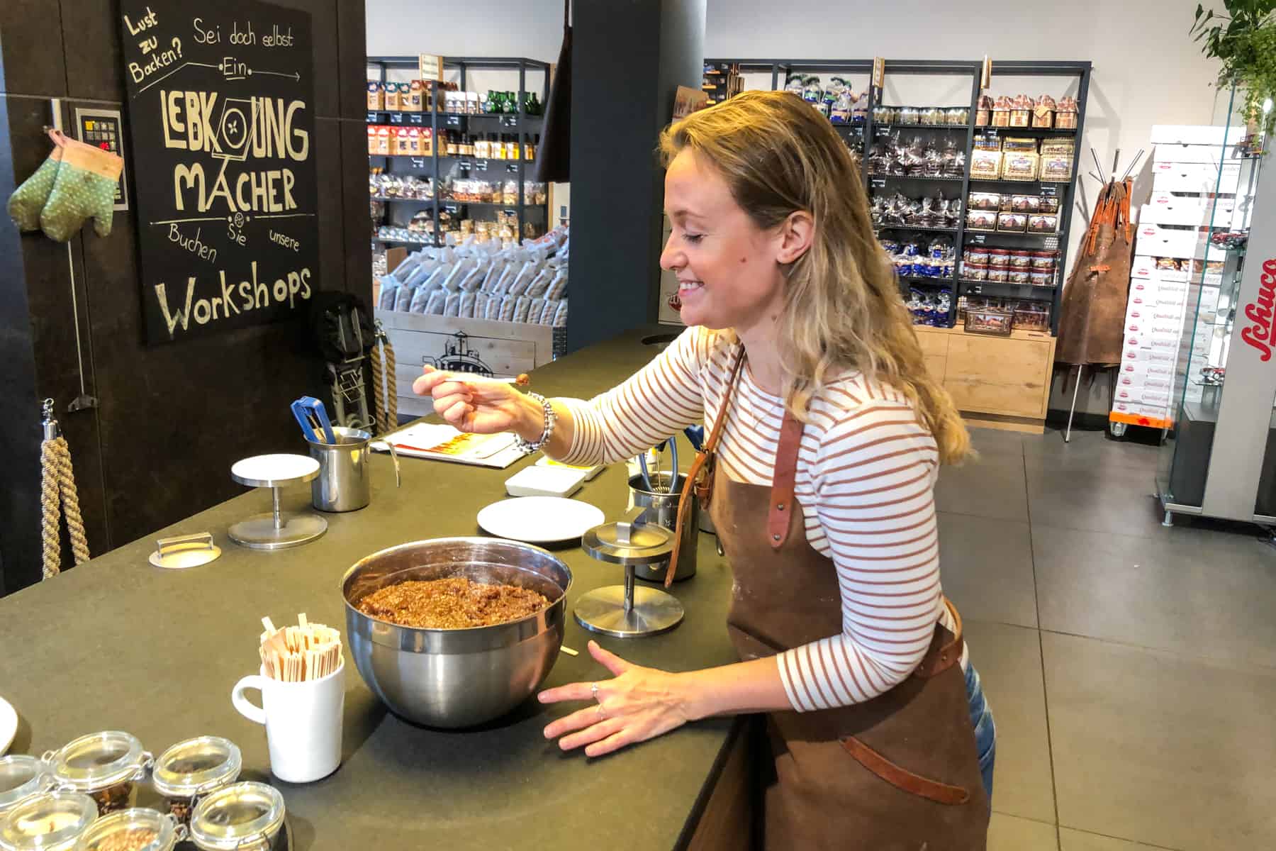 Los mejores lugares para Comer y Beber en Nuremberg Una mujer con un top a rayas y un delantal de cuero marrón toma una cucharada de la mezcla de pan de jengibre de Nuremberg de un tazón grande de metal y está a punto de comerlo.