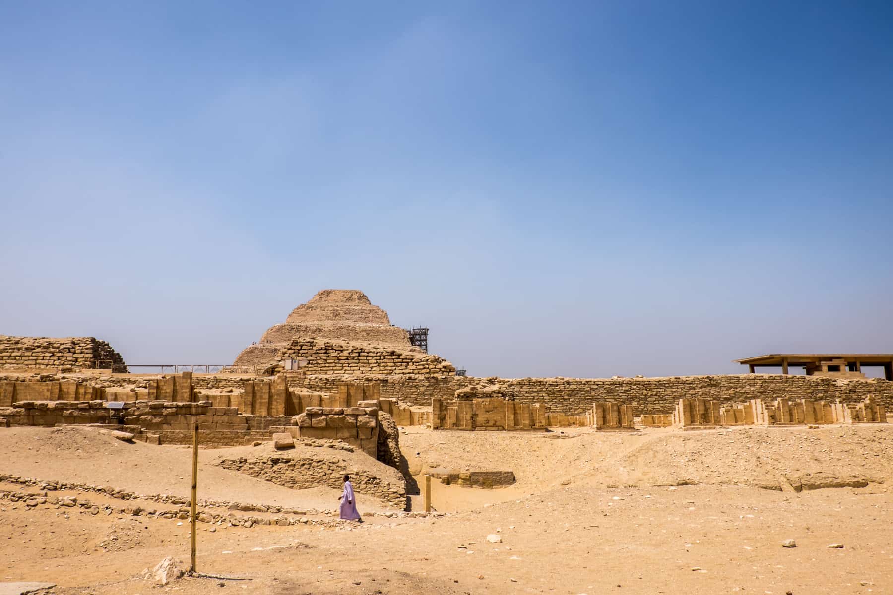 An Egyptian man in a purple robe walks in the sand in front of the step pyramid in Saqqara, Egypt