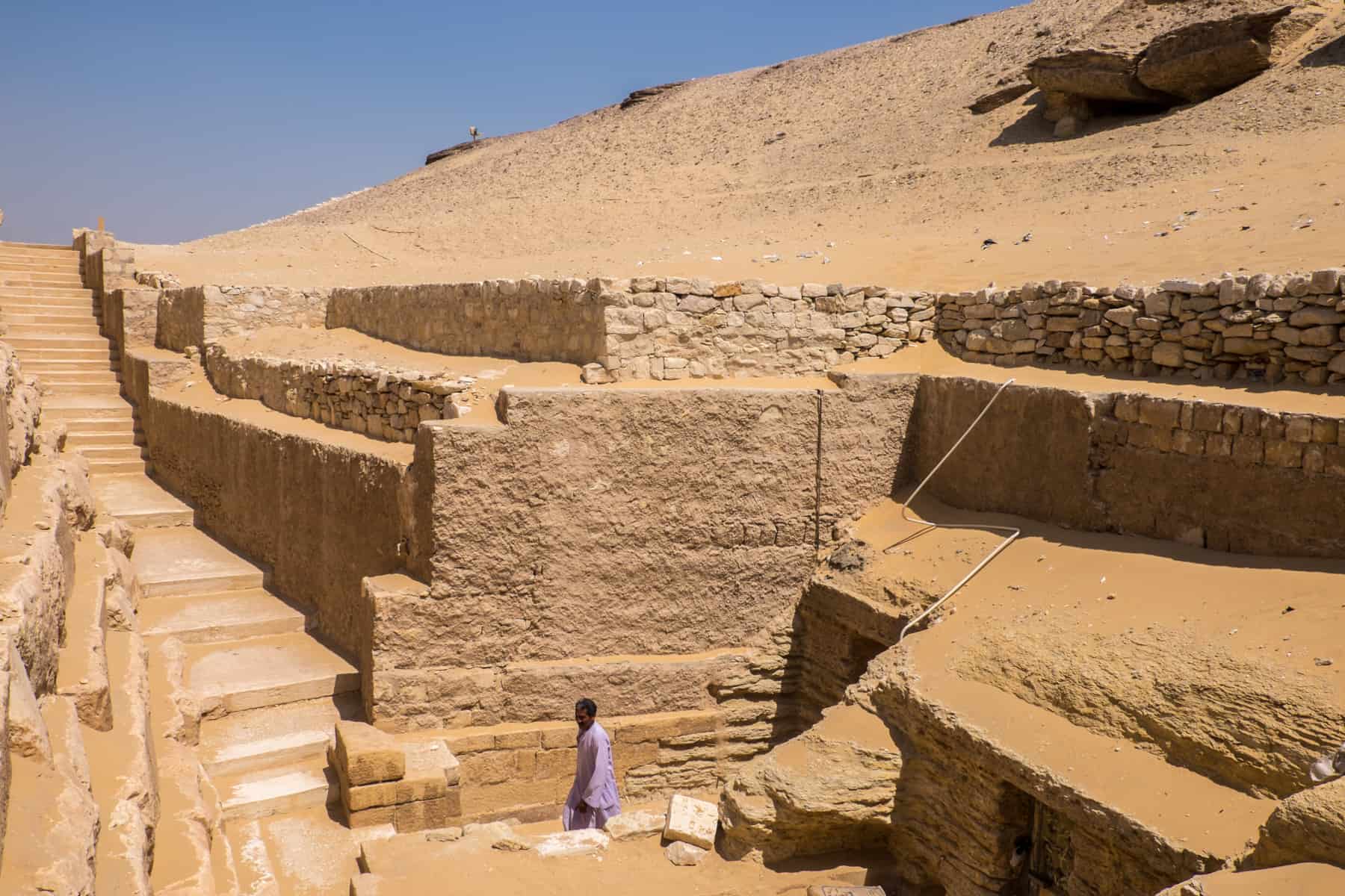 An Egyption guard in lilac robe exits the doorway of a sandy, tiered tomb complex in Saqqara, Egypt