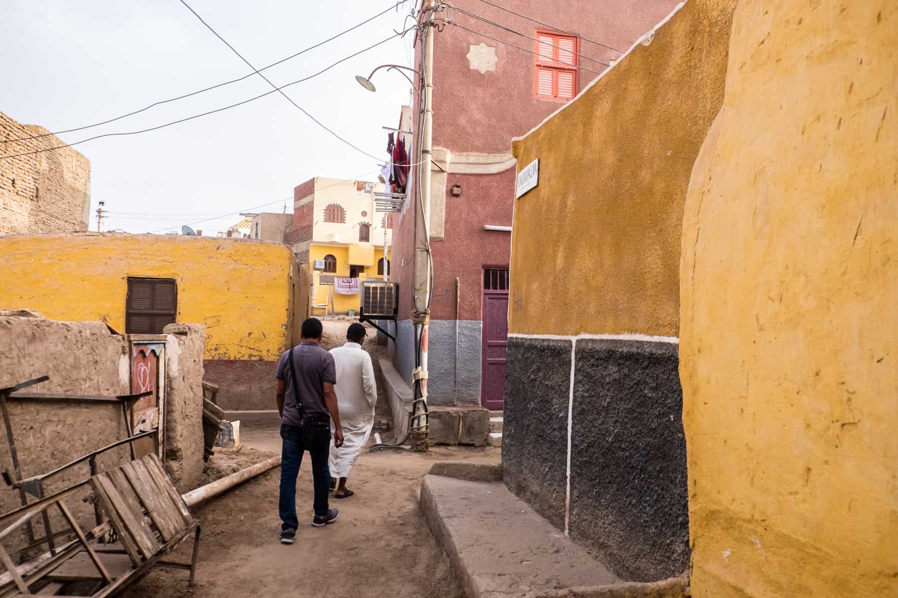 An Egyptian guide and Nubian man walk through the pink and yellow painted walls of a traditional Nubian village in Aswan, Egypt
