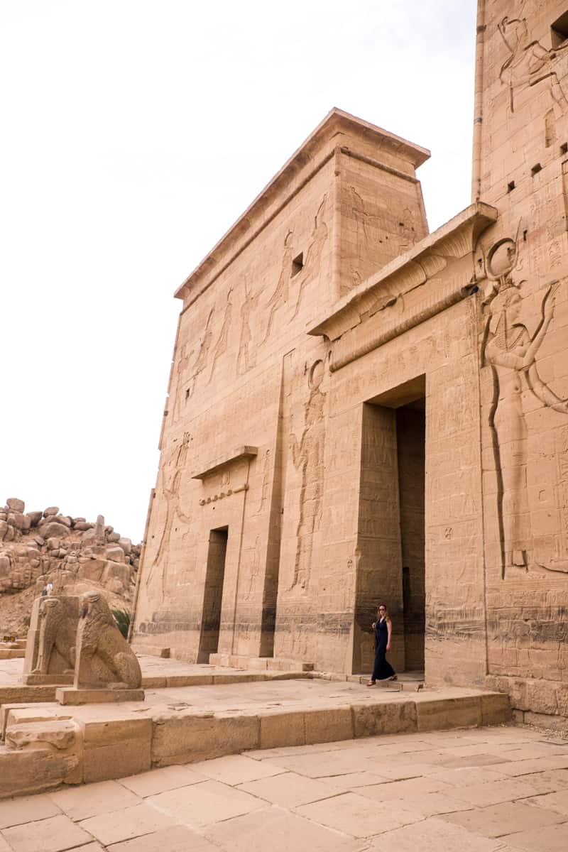 A woman in a blue dress walks out of the entrance to the grand, rectangular Philae Temple in Aswan, Egypt