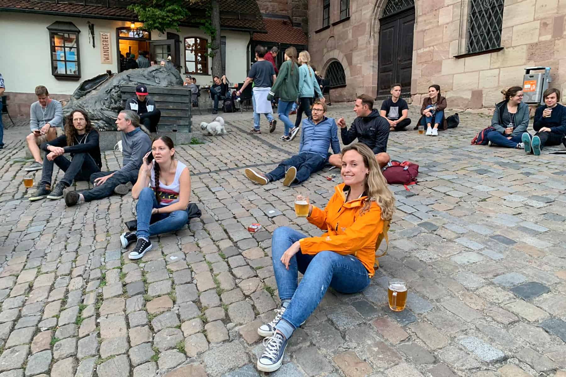 A woman in an orange jacket and jeans drinks a large beer while sitting on the cobblestoned streets of Nuremberg Old Town