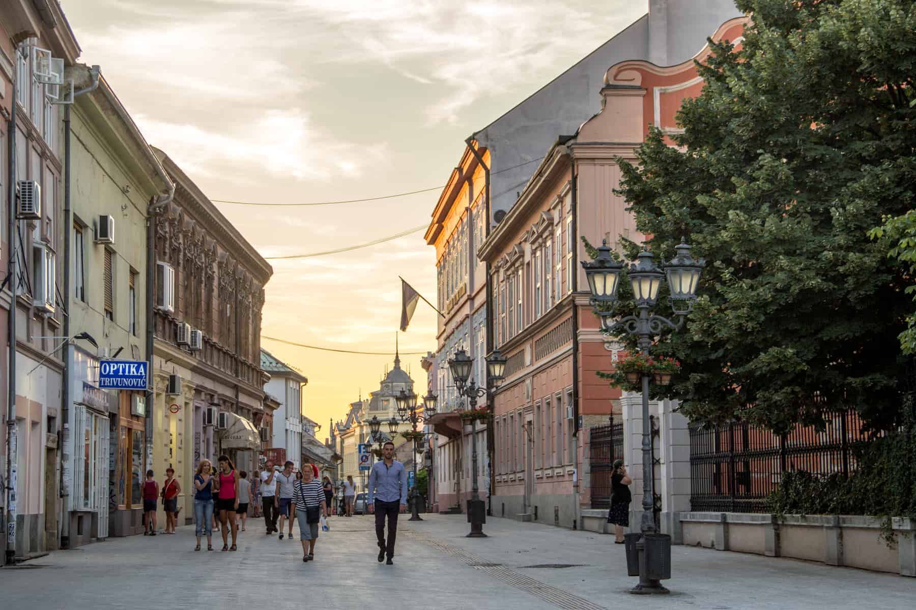 People walking down a pedestrian street in Nove Sad, Serbia, lined with pastel coloured buildings, glowing from the sunset in the background. 