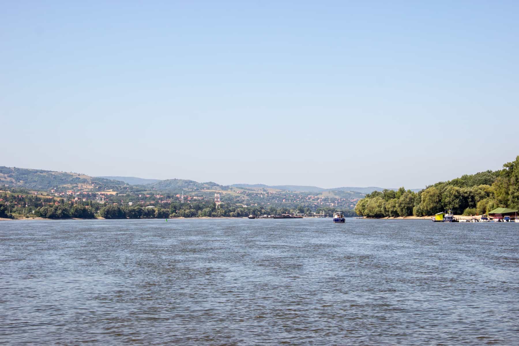 The wide expanse of the Danube River's blue water in Serbia. In the distance is a boat, low rolling hills covered in houses and to the right, a yellow sand island. 