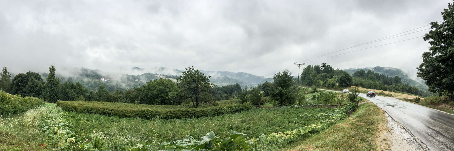 A car on a the end of a road curving around a hill backed forest. 