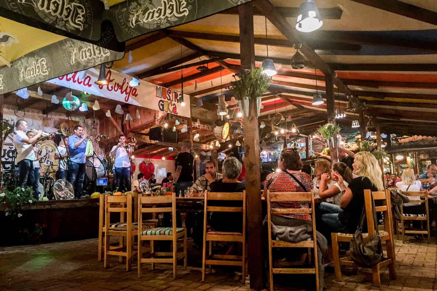 A band of trumpet players plays to an audience sitting around large wood tables. A typical scene from the Trumpet Festival in Serbia. 