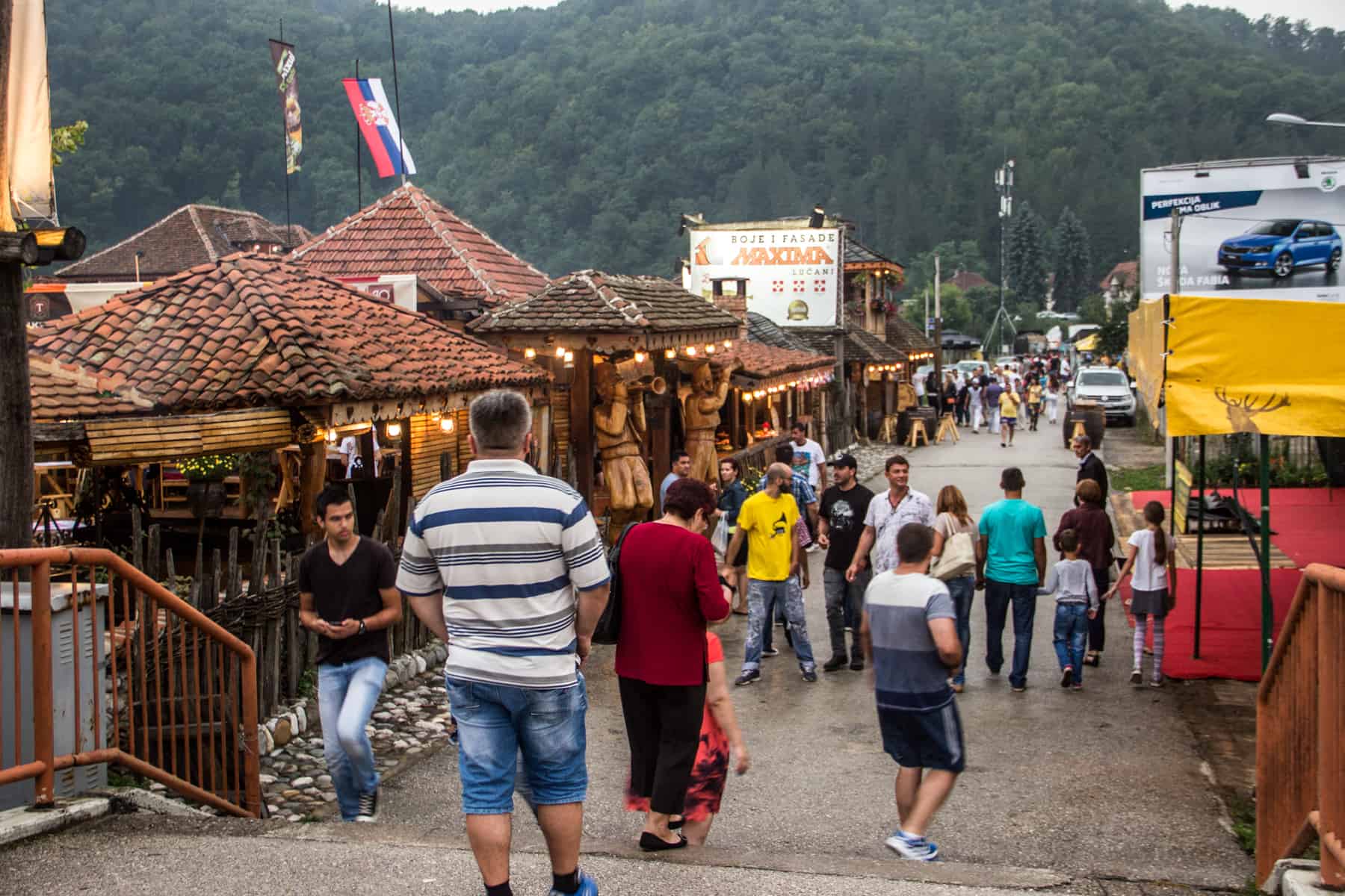 People walking in both direction down a street with wooden houses with stone roofs, in the Serbian village of Guca.