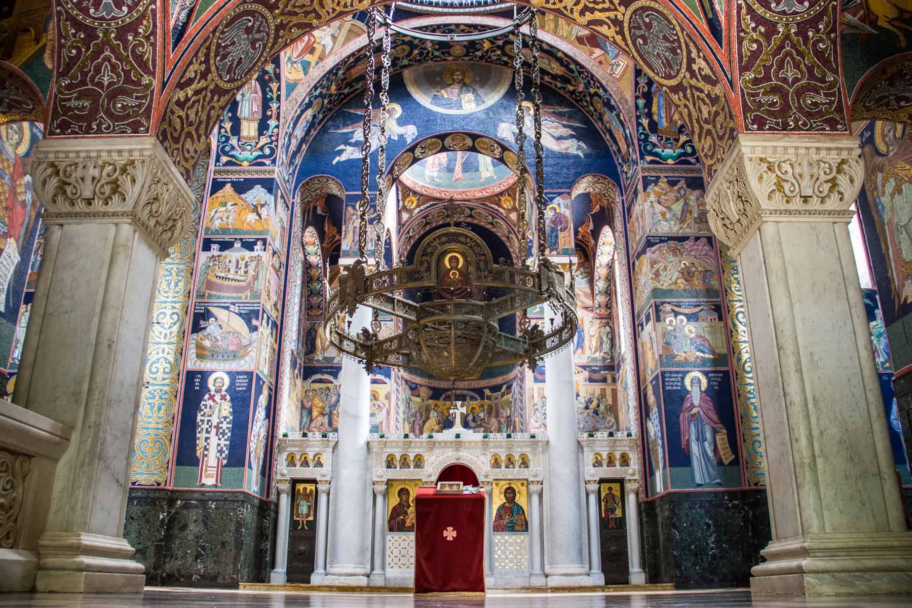 The interior of the St. Georges Church in Serbia, decorated with mosaic pictures mainly in a deep blue and gold. A large bronze chandelier hangs in the centre. 