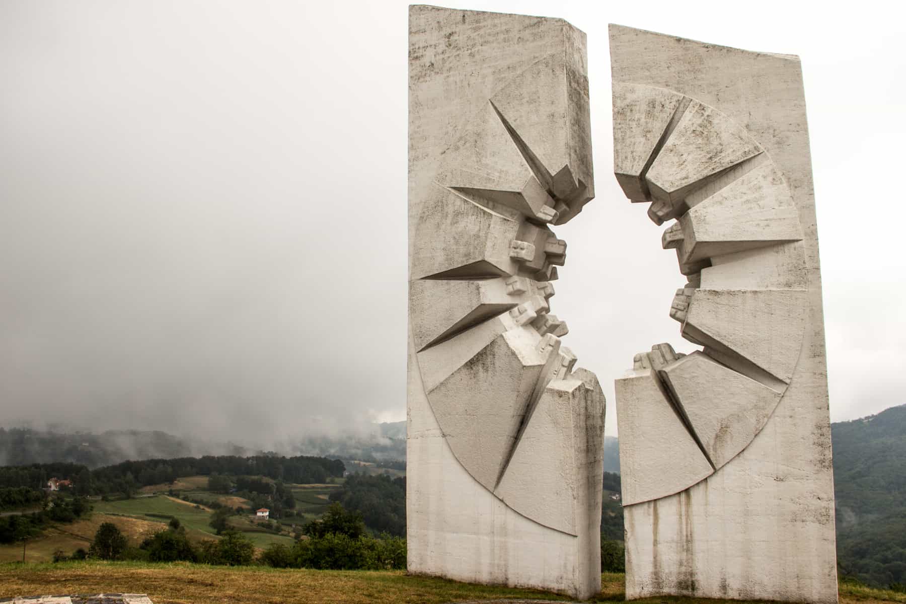 A large white stone, carved to look like the shattering from a bullet, overlooks a green mountainous valley.