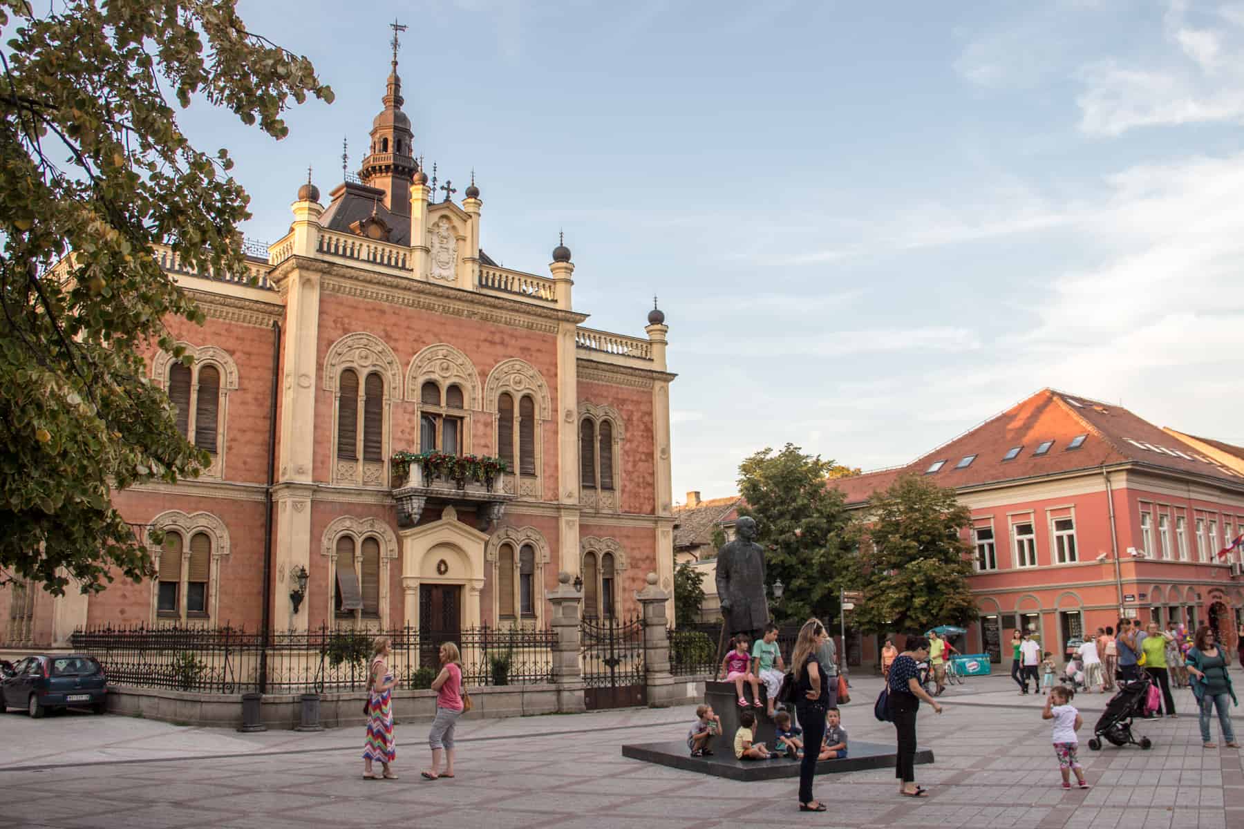 People gather around a small, black statue outside an opulent, mansion-like orange and yellow building in Novi Sad, Serbia. 