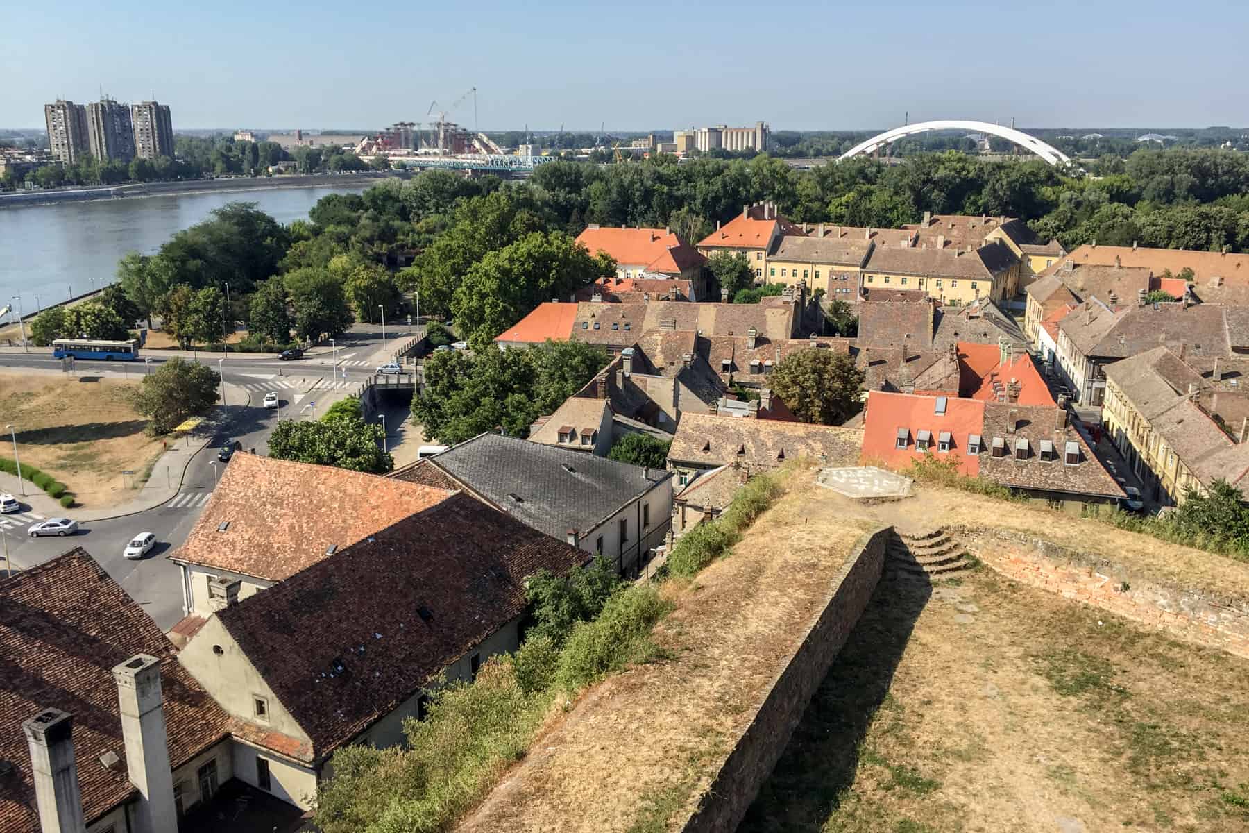 Elevated view from the corner stone of a Fortress in Novi Sad, overlooking a group of brown and orange roof houses, the river, a white arch and skyscrapers in the distance. 