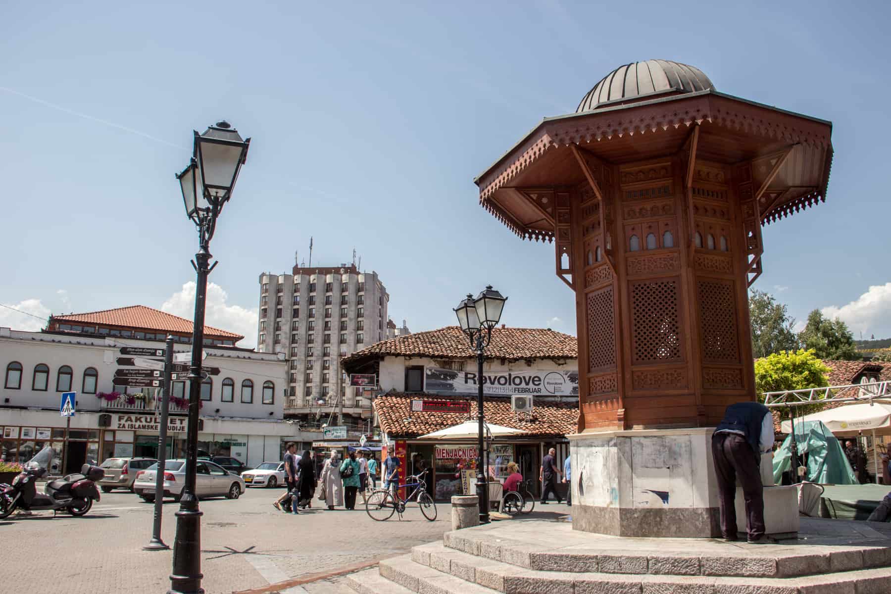 A man drinks water from an ochre red and white marble structure that stands with a public square surrounded by white buildings and orange rooftops. 