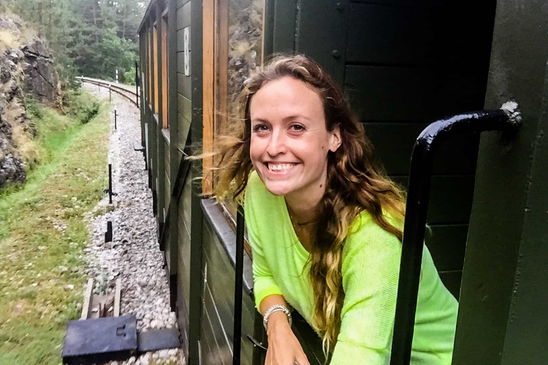 A woman smiles while leaning out of a green, wooden train carriage to a backdrop of forest. 