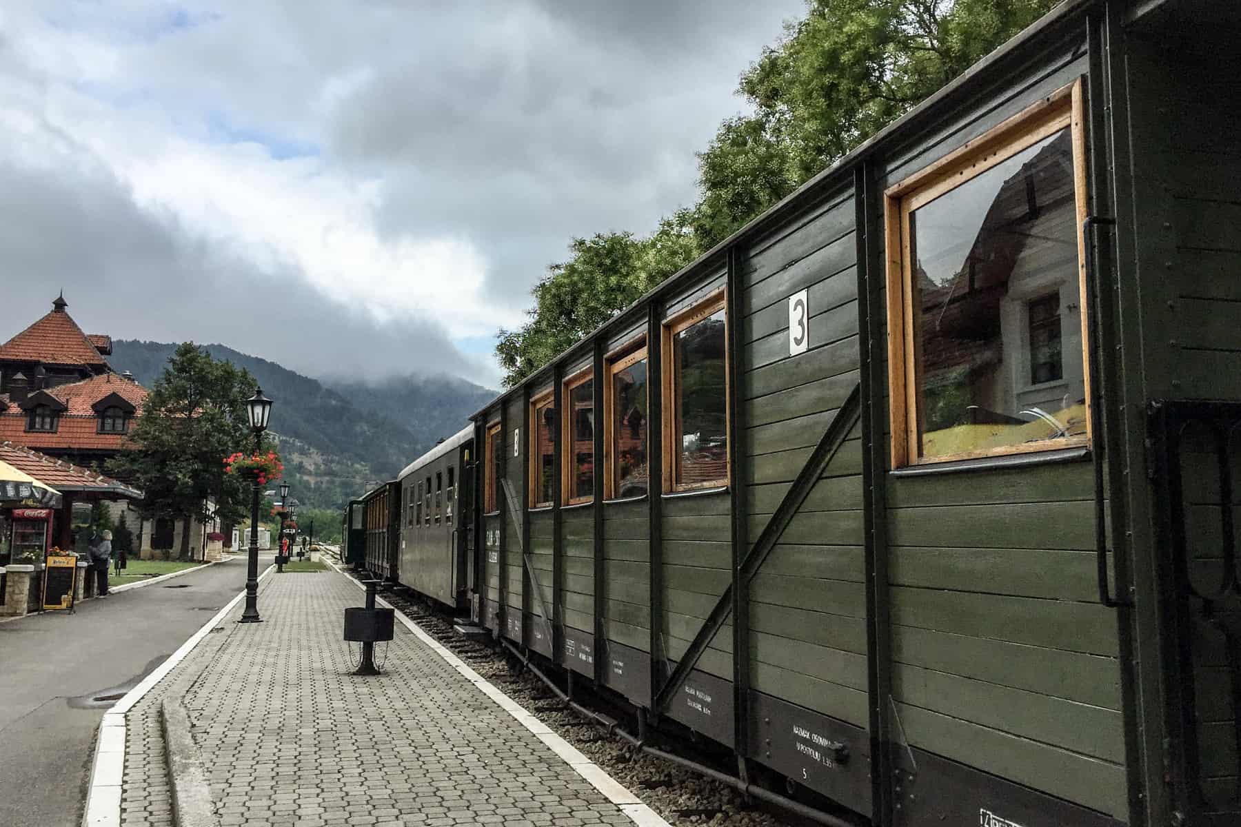 A dark green wood panelled train - The Sargan Eight Railway in Serbia - at the station platform with orange roofed buildings, in the direction of the mountains.
